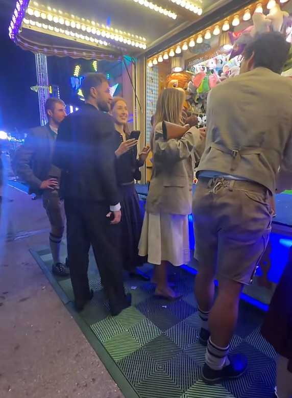 Mehrere Menschen in Tracht stehen an einem beleuchteten Jahrmarkt-Stand mit Plüschtieren.