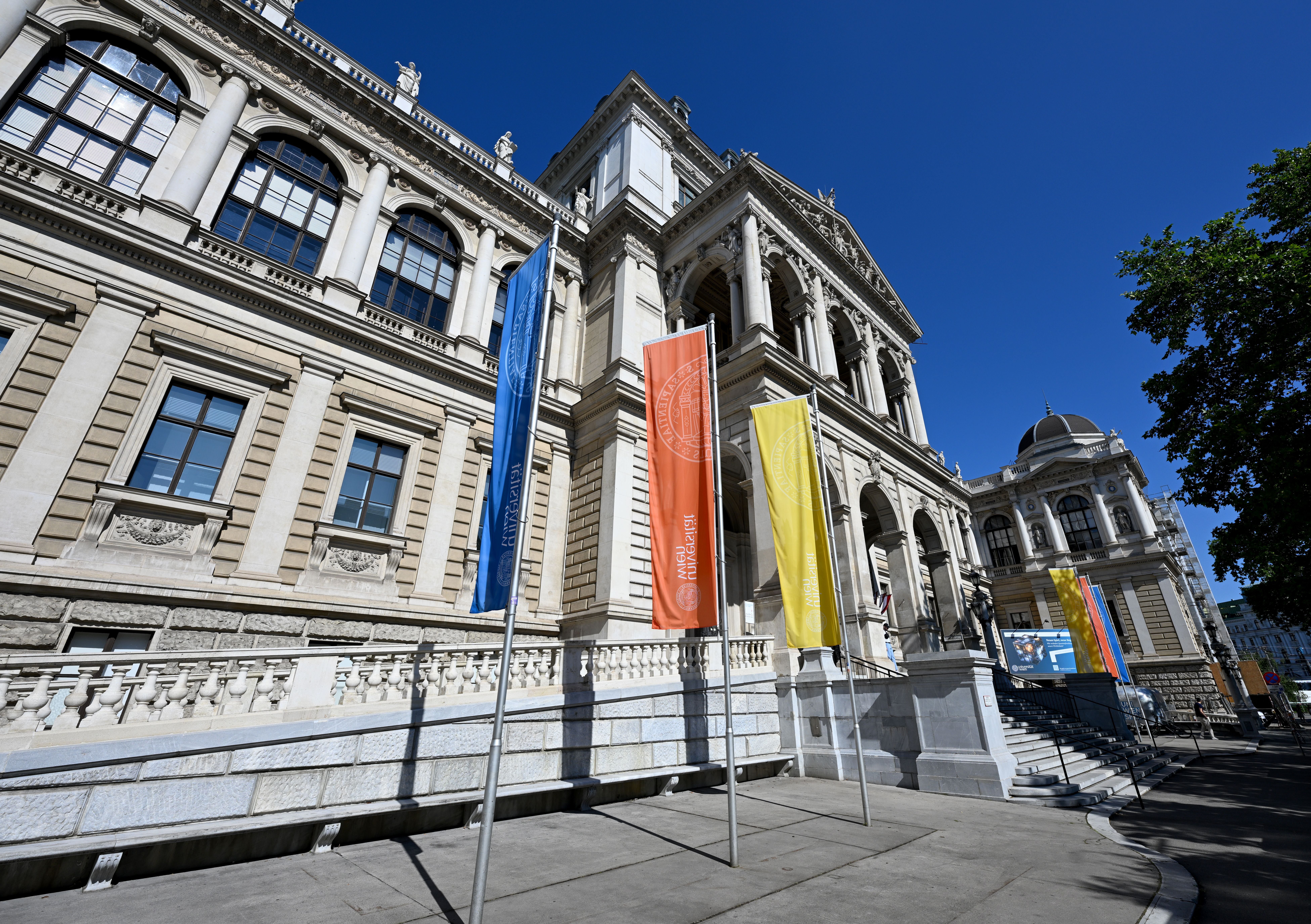 Fassade des Hauptgebäudes der Universität Wien mit bunten Fahnen und blauem Himmel.