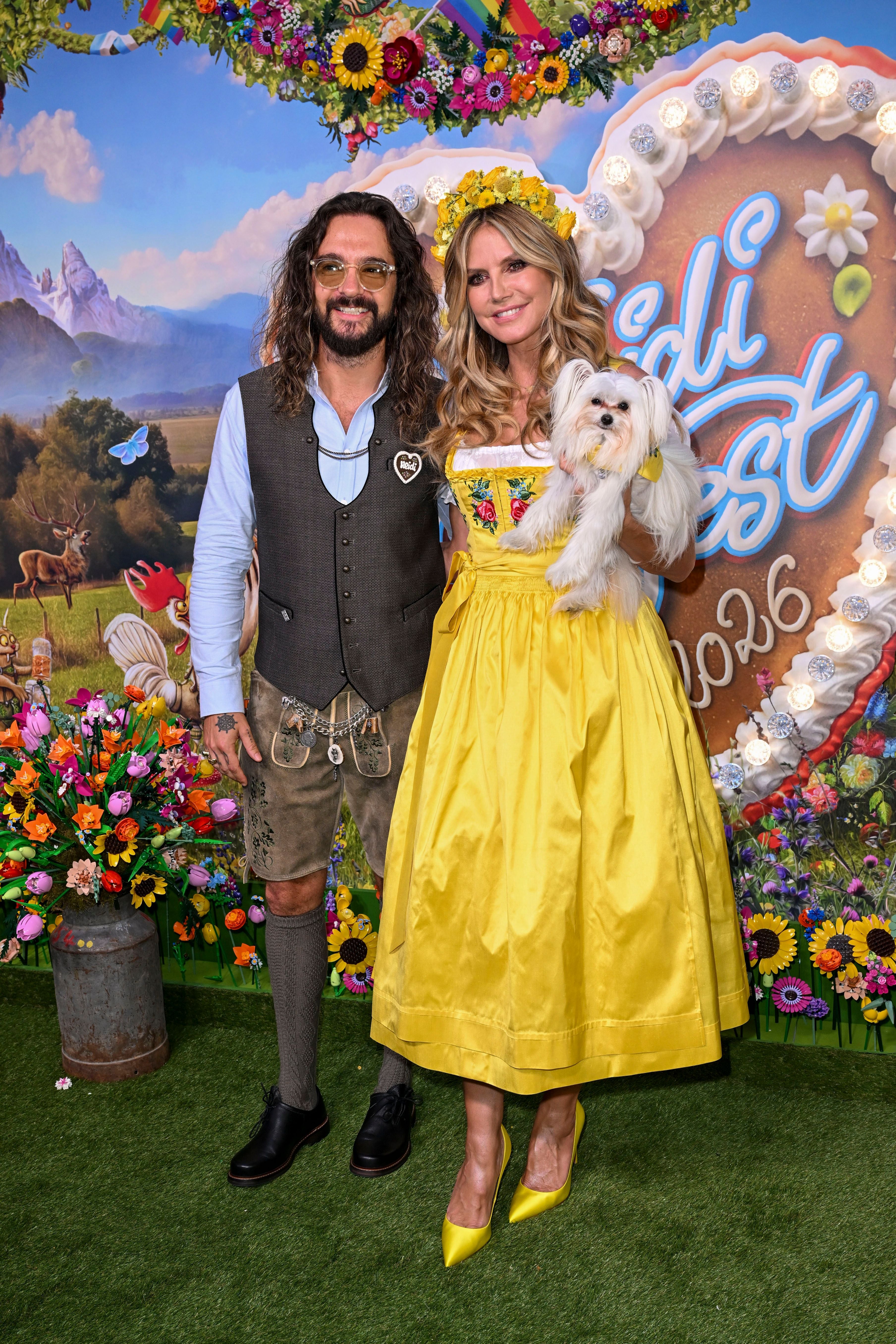 Ein Mann in Tracht und eine Frau im gelben Dirndl mit Blumenkranz und Hund posieren vor Blumenwand.