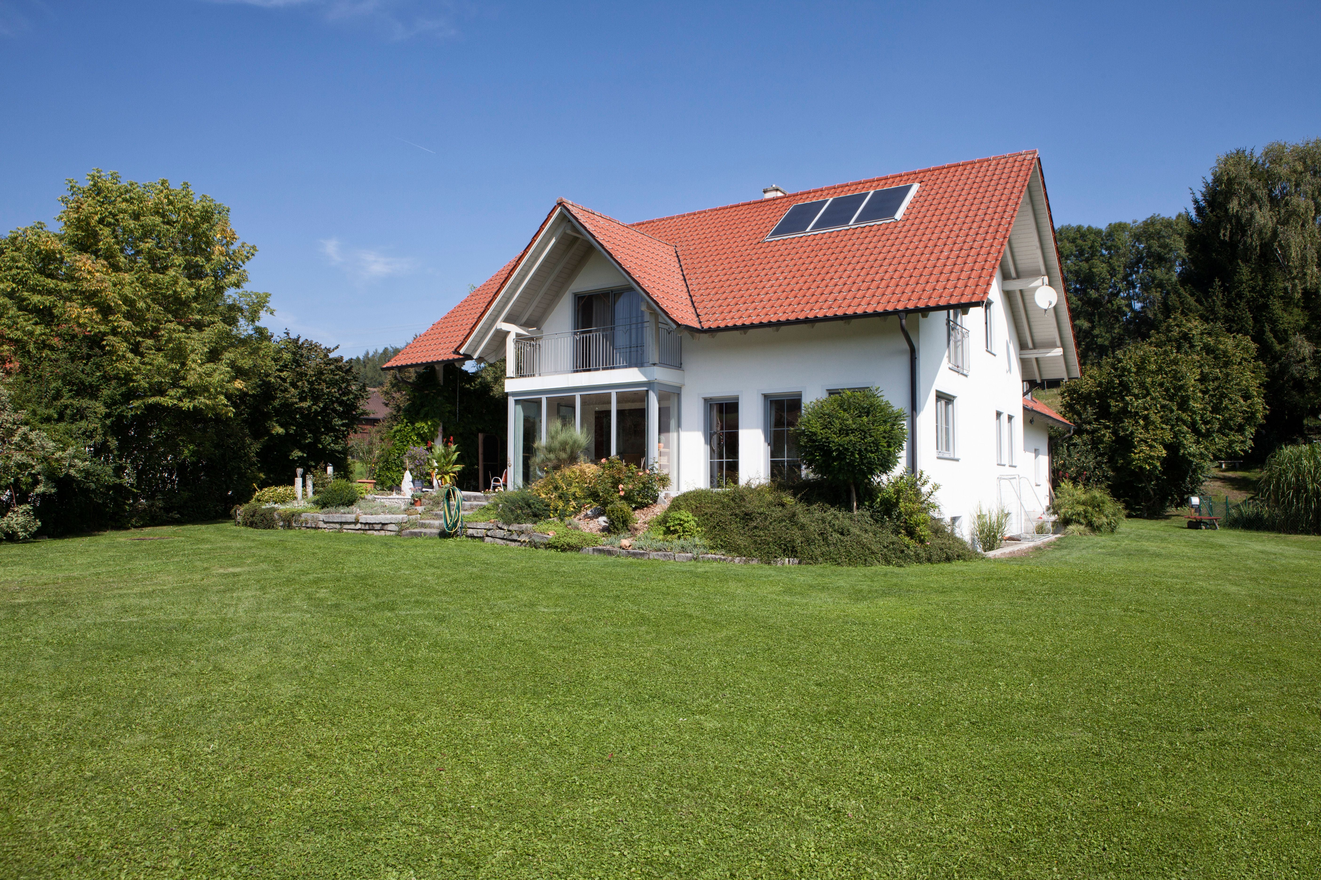 Modernes Einfamilienhaus mit rotem Dach und Balkon in großem grünen Garten bei Sonnenschein.
