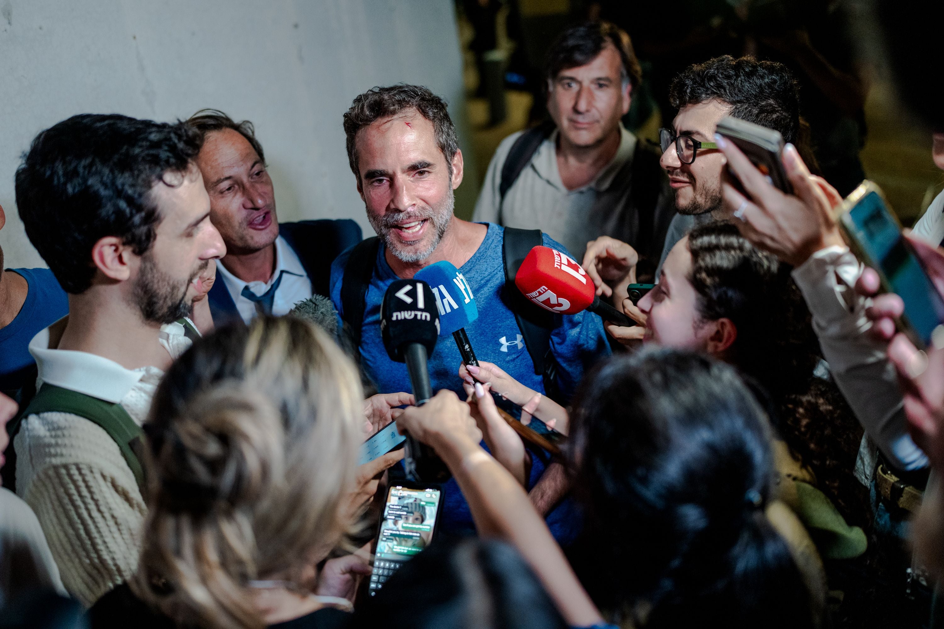 Mann in blauem Shirt spricht am Flughafen umringt von Reportern mit Mikros und Handys.
