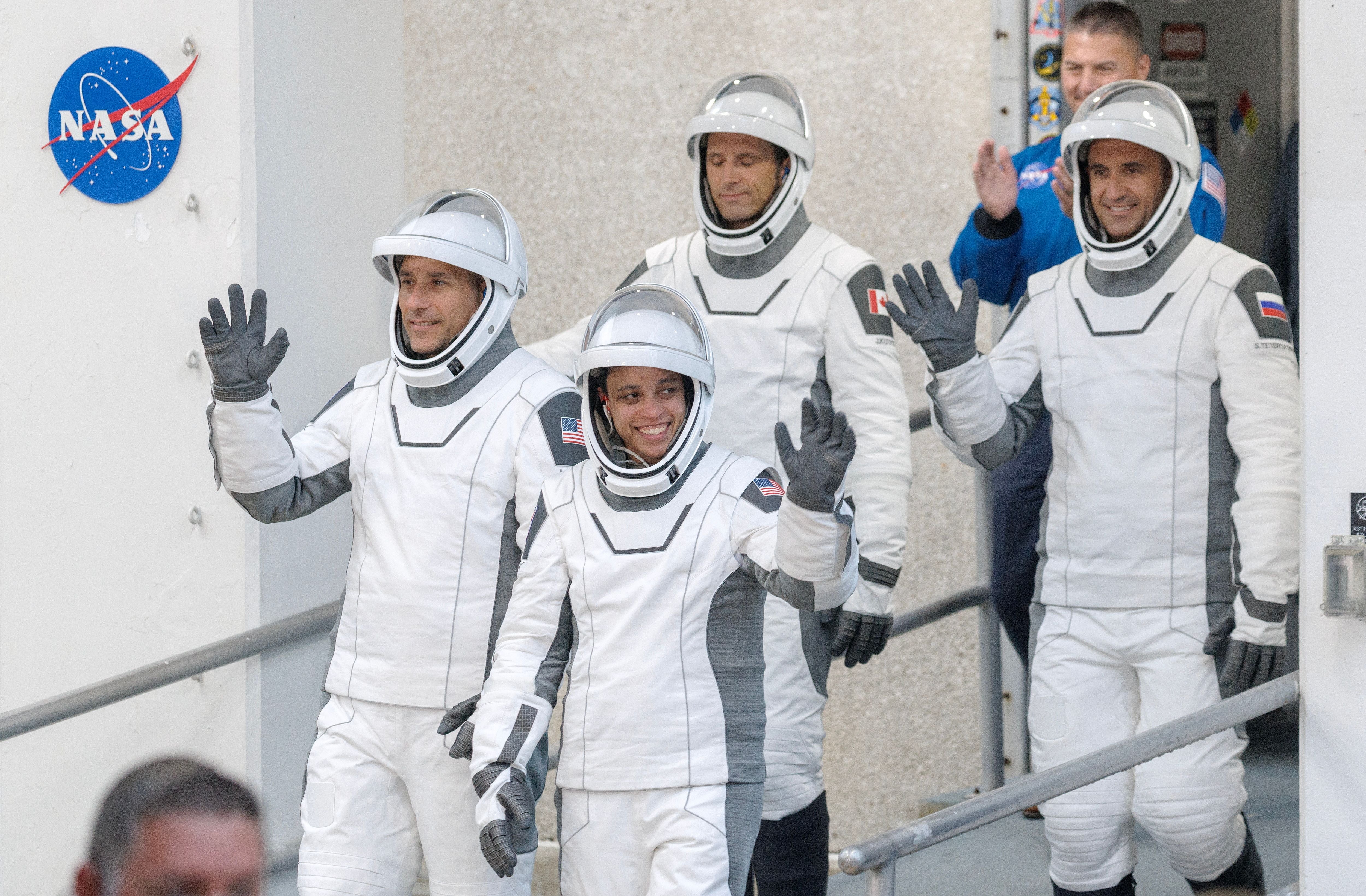 Vier Astronauten in Raumanzügen winken beim Verlassen eines Gebäudes mit NASA-Logo.