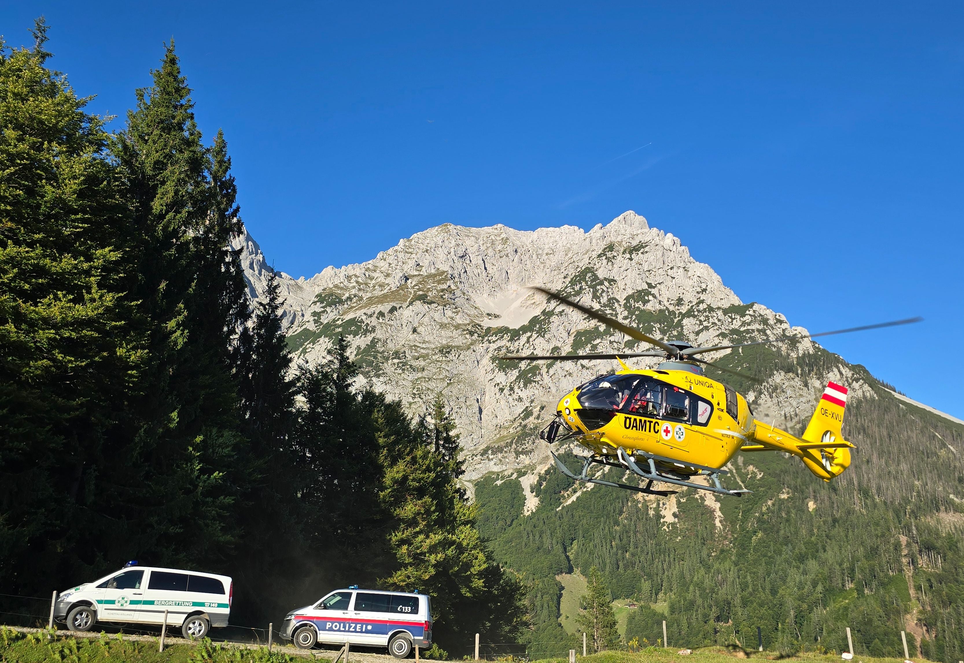 Ein gelber Rettungshubschrauber fliegt vor Gebirgslandschaft, unten stehen Polizei- und Einsatzfahrzeug.
