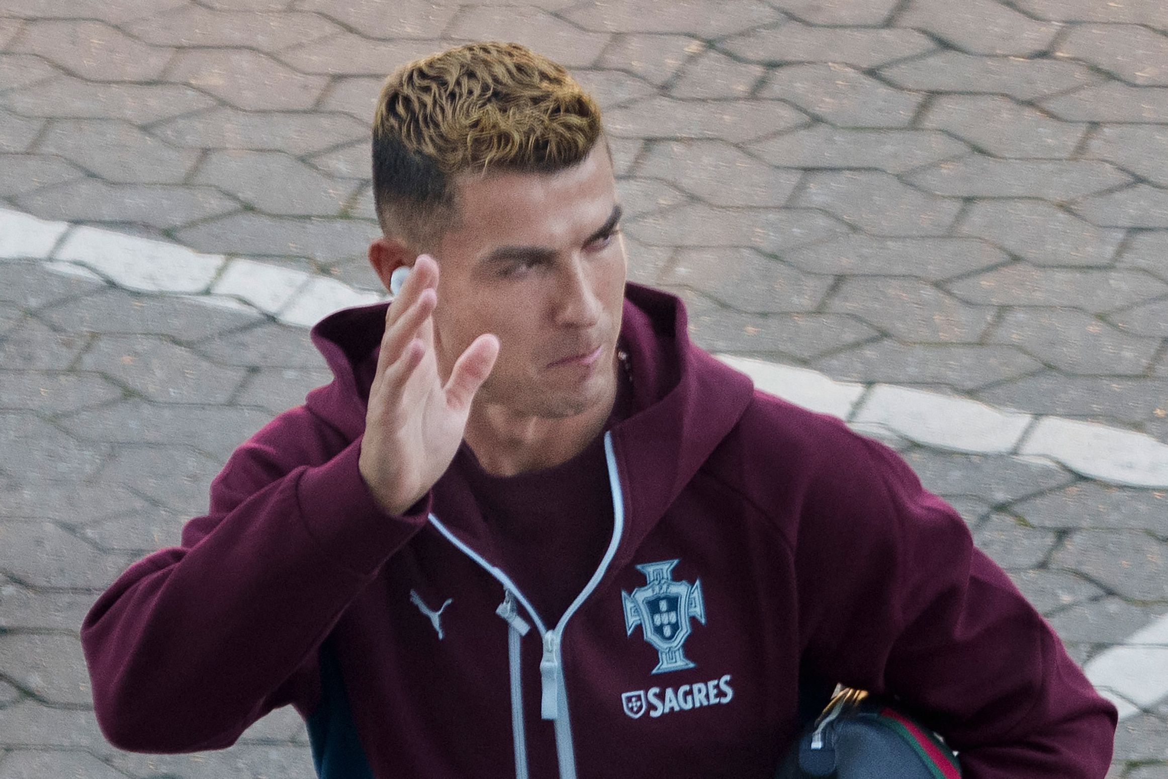 Ein Mann in burgunder Trainingsjacke winkt, Hintergrund ist Kopfsteinpflaster.