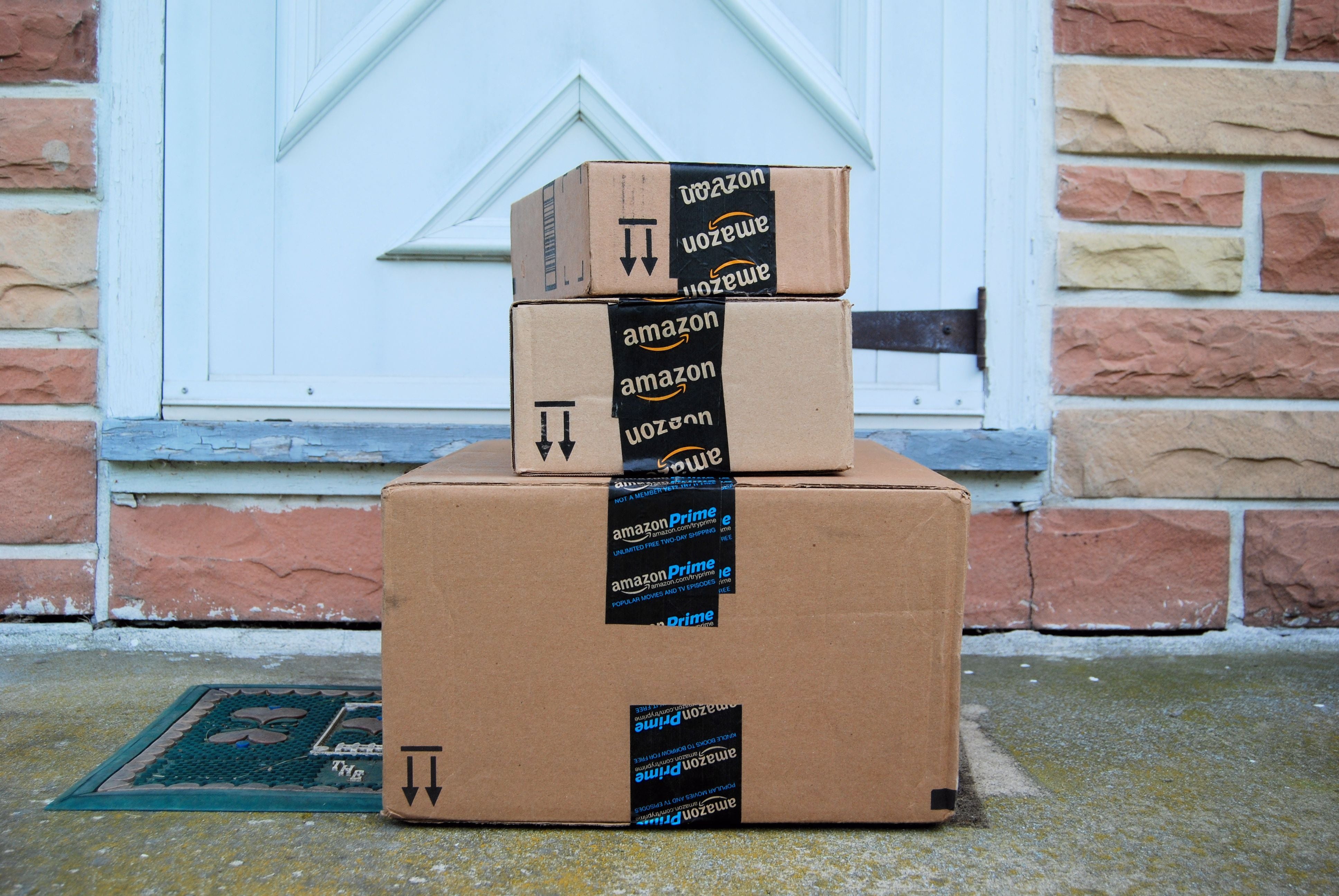 Drei Amazon-Pakete stehen gestapelt vor einer Haustür auf einer Fußmatte.