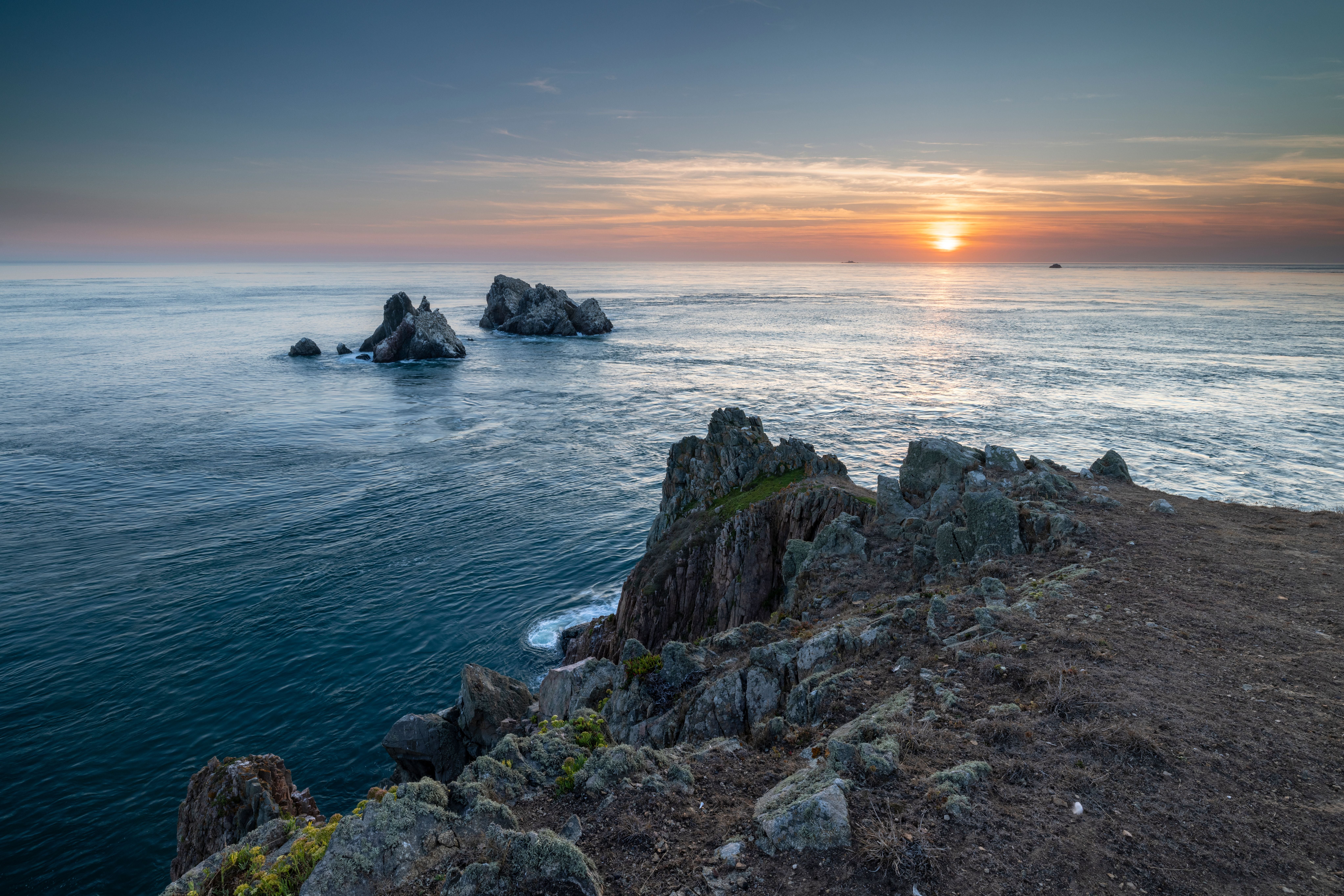 Felsige Küste von Alderney mit rauer See und Sonnenuntergang am Horizont.