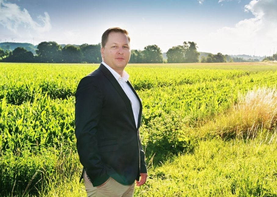 Ein Mann im Anzug steht auf einem Feld vor einer grünen Landschaft und blauem Himmel.