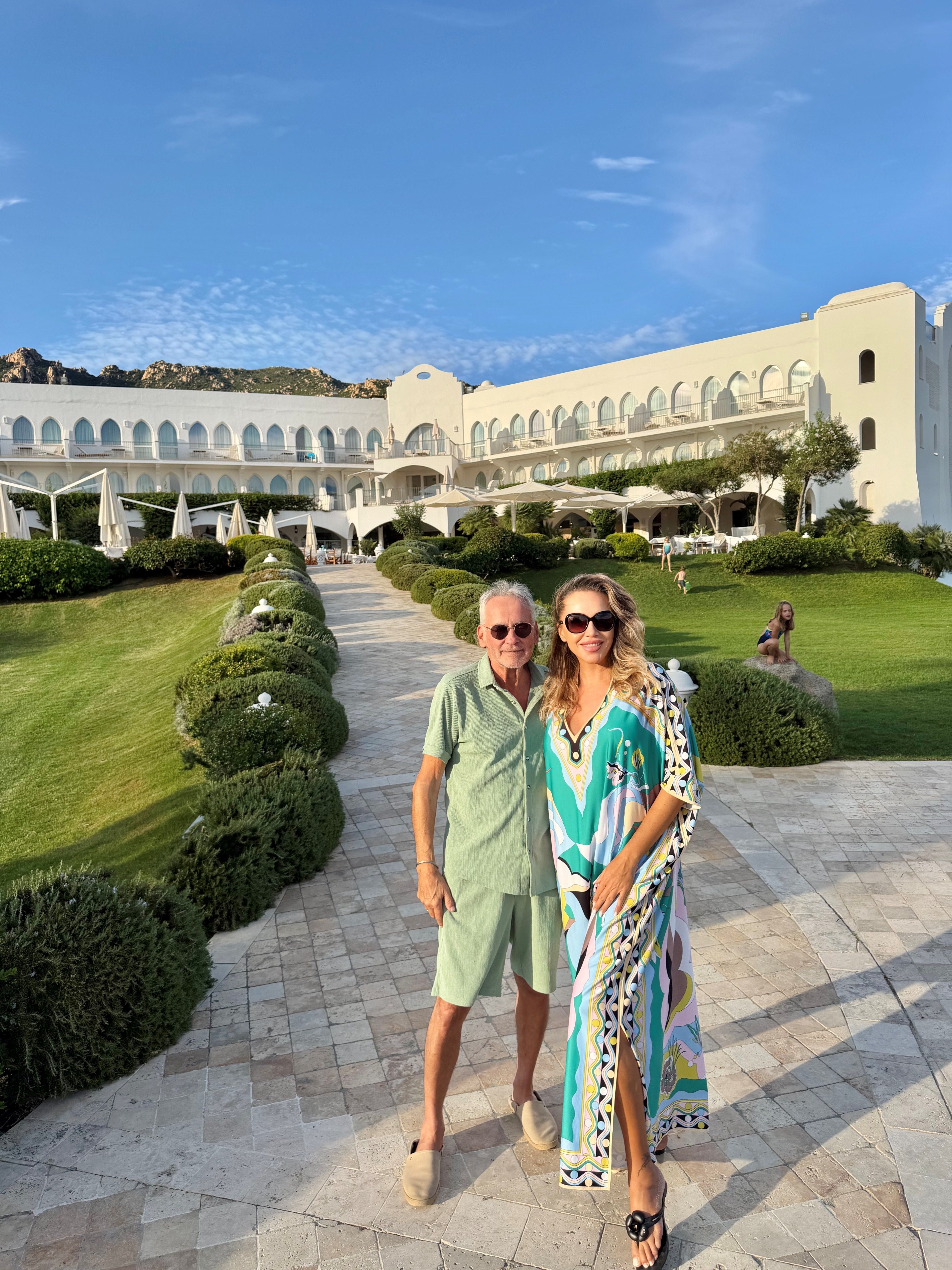 Ein Mann und eine Frau posieren vor einem eleganten, weißen Hotelgebäude im Sonnenschein.