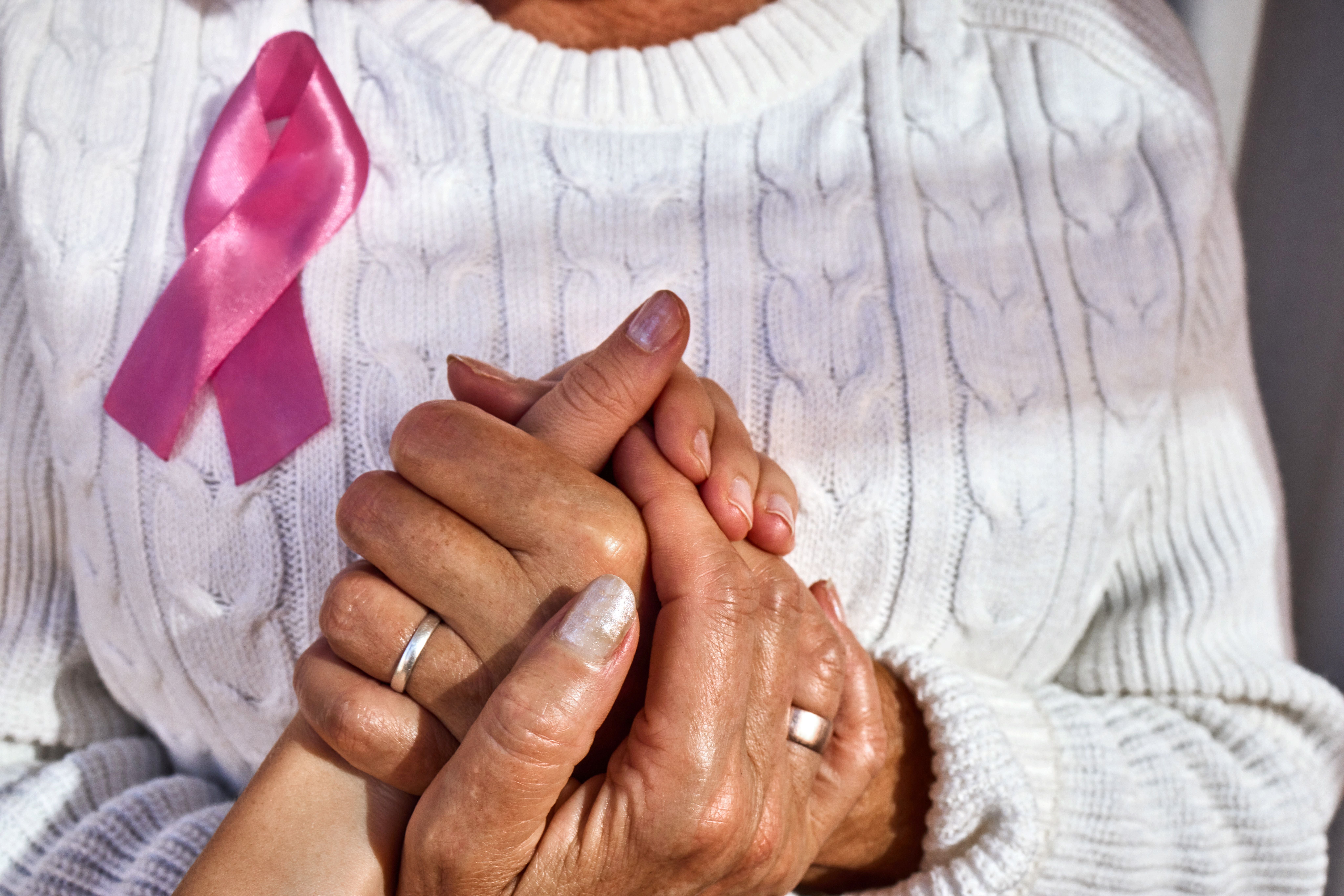 Zwei Menschen halten Hände, eine Person trägt eine rosa Schleife am weißen Pullover.