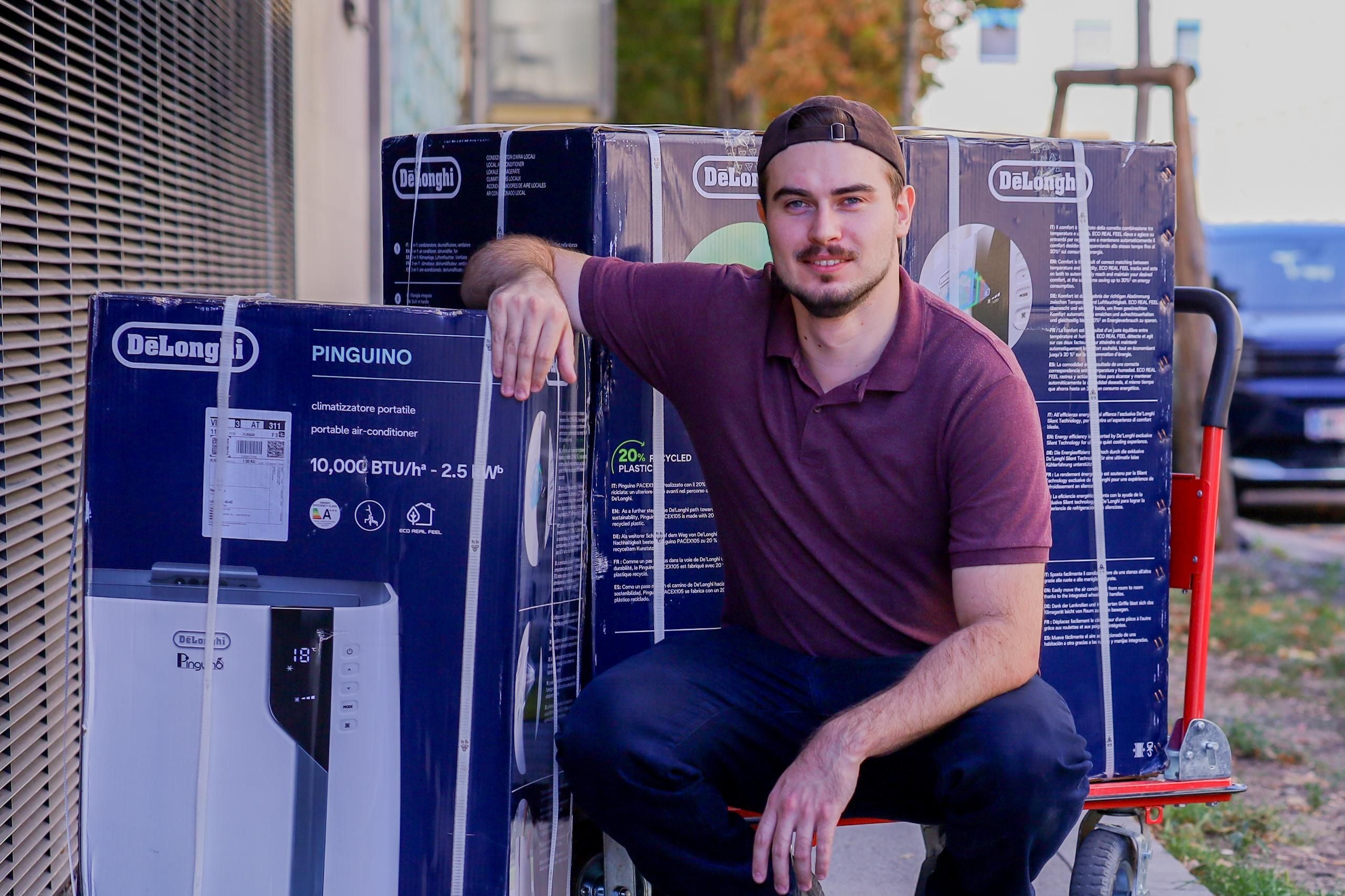 Ein Mann sitzt neben mehreren großen De'Longhi-Kartons auf einer Sackkarre auf dem Gehweg.
