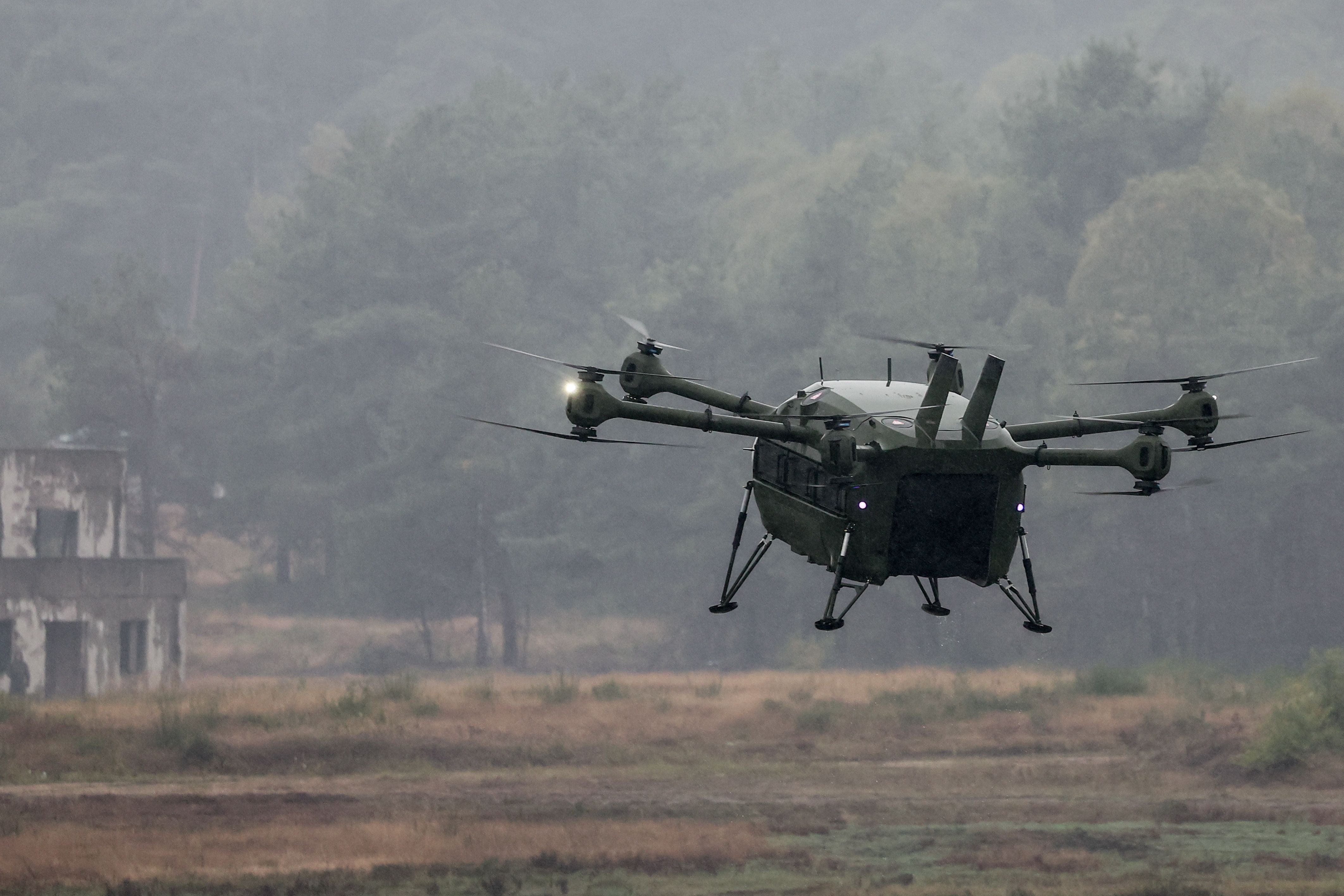 Große militärische Drohne fliegt über ein Feld mit Bäumen und einem verlassenen Gebäude im Hintergrund.