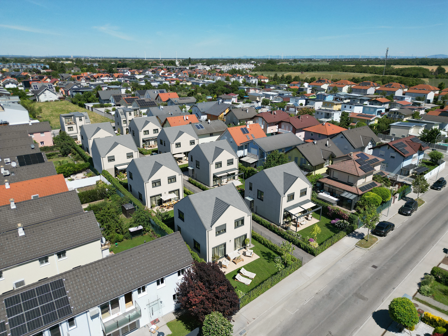 Luftaufnahme eines Wohngebiets mit Reihenhäusern, Gärten und Straße bei sonnigem Wetter.