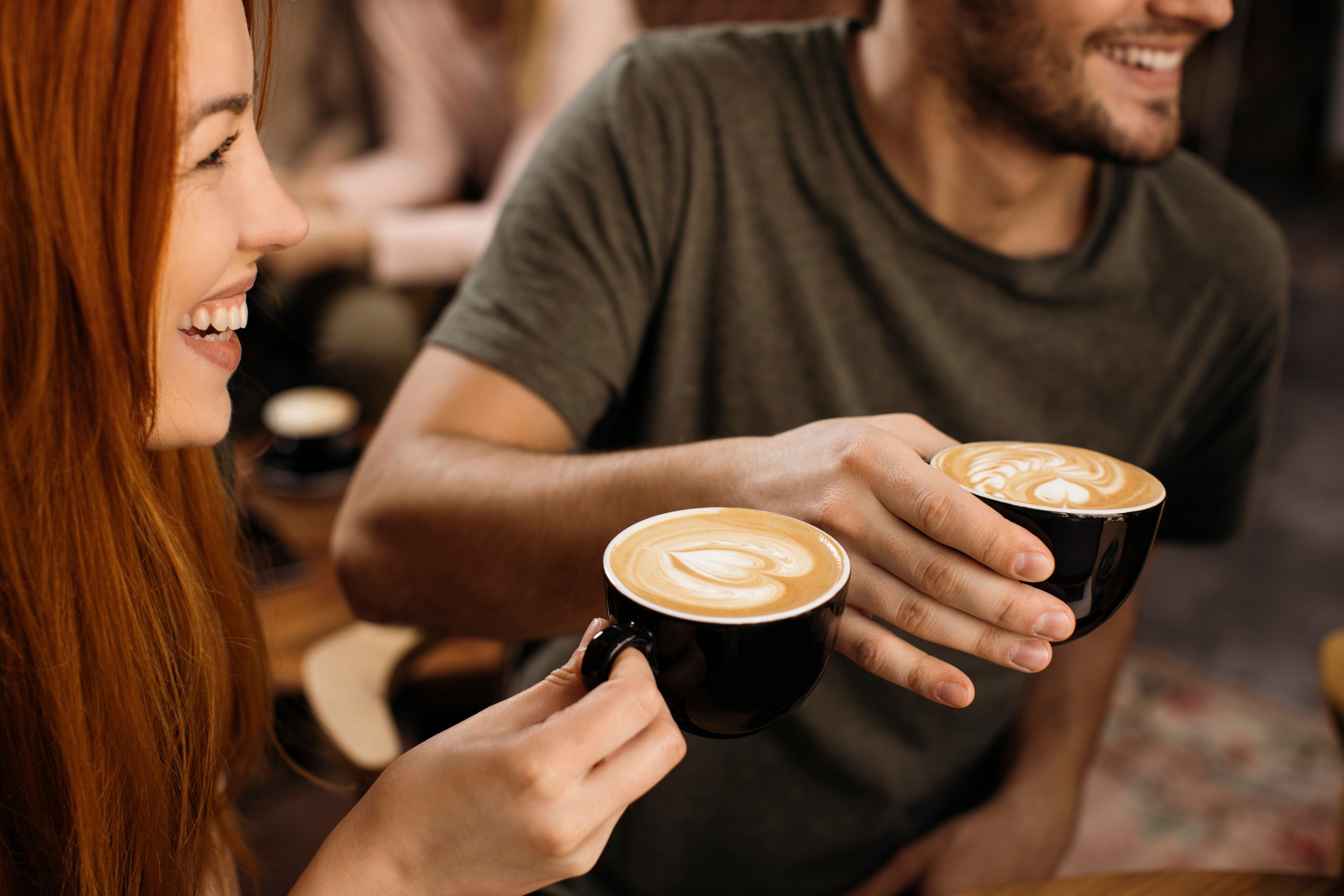 Zwei lachende Freunde stoßen mit Tassen Cappuccino mit Latte Art an.