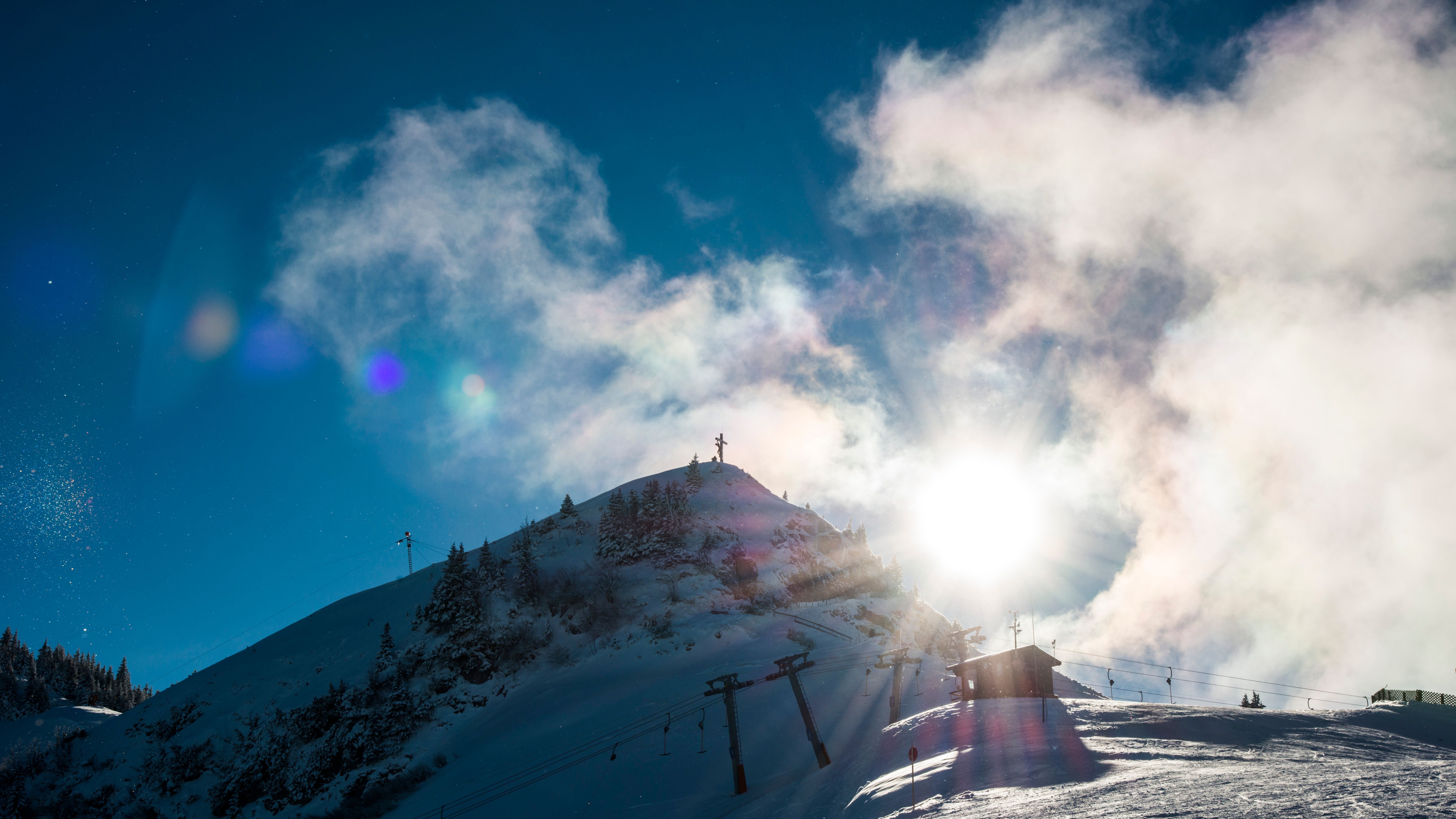 Verschneiter Berg mit Gipfelkreuz, Seilbahn und strahlender Sonne im Hintergrund.