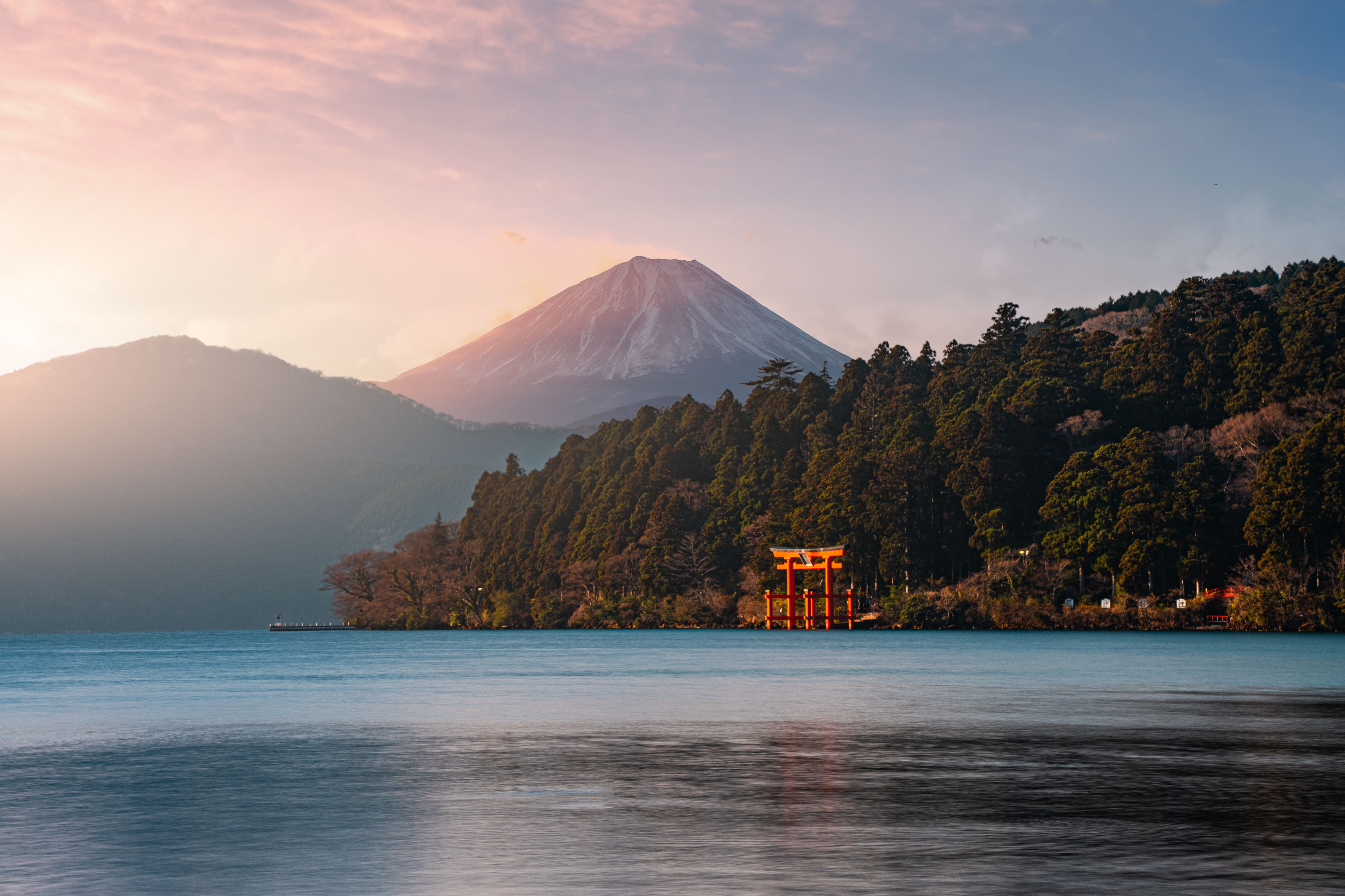 Rotes Torii-Tor am See vor bewaldeter Küste, im Hintergrund der schneebedeckte Berg Fuji.