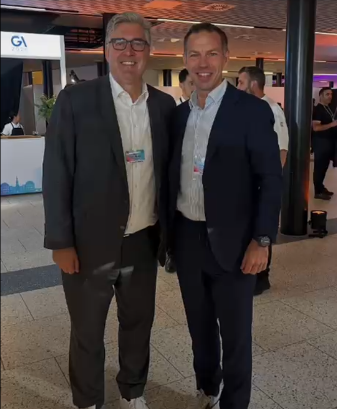 Axel Hellmann und Christian Peschek stehen lächelnd nebeneinander in Anzügen im Foyer einer Veranstaltung.