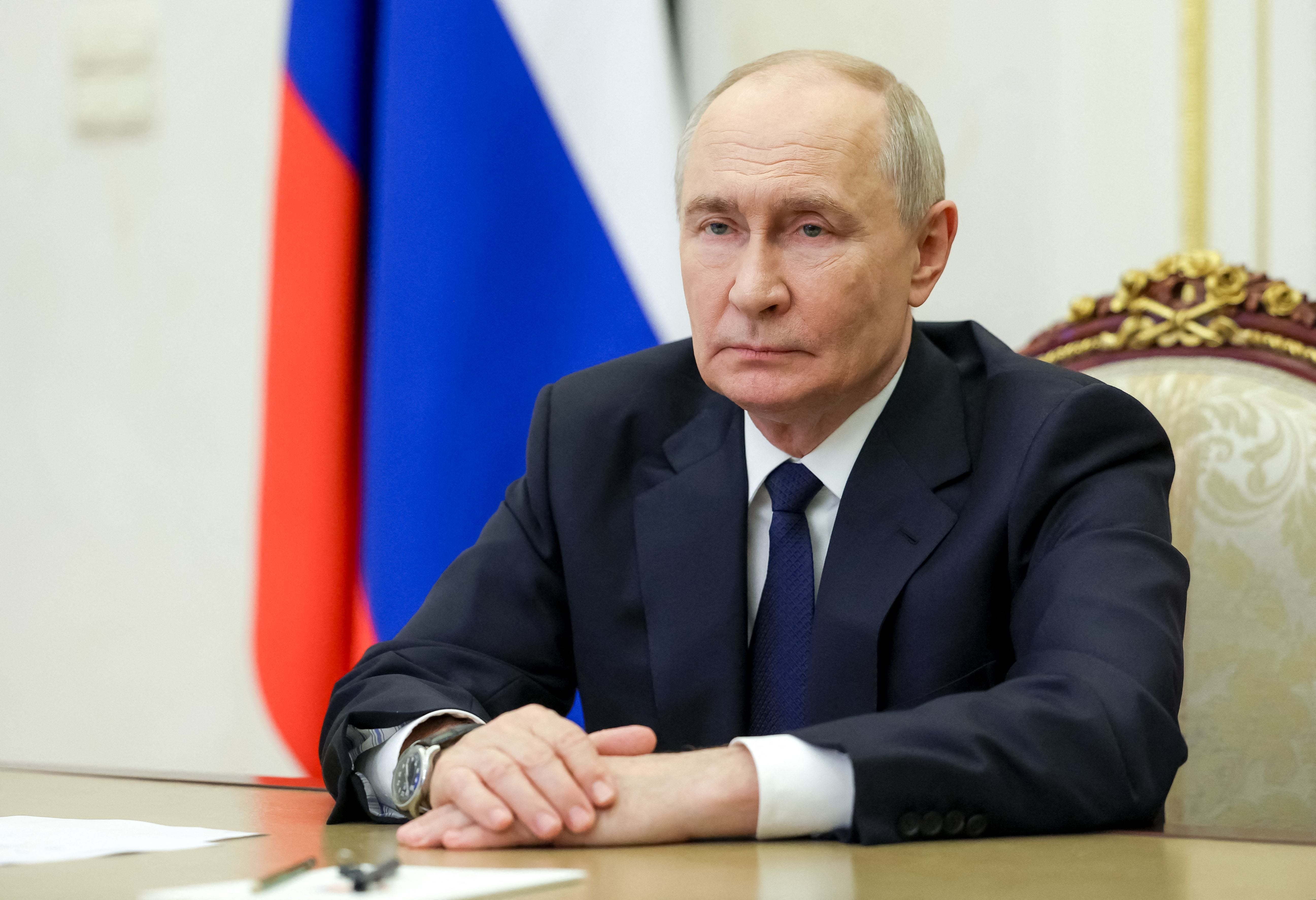 Ein Mann im Anzug sitzt an einem Tisch vor einer russischen Flagge und schaut ernst.
