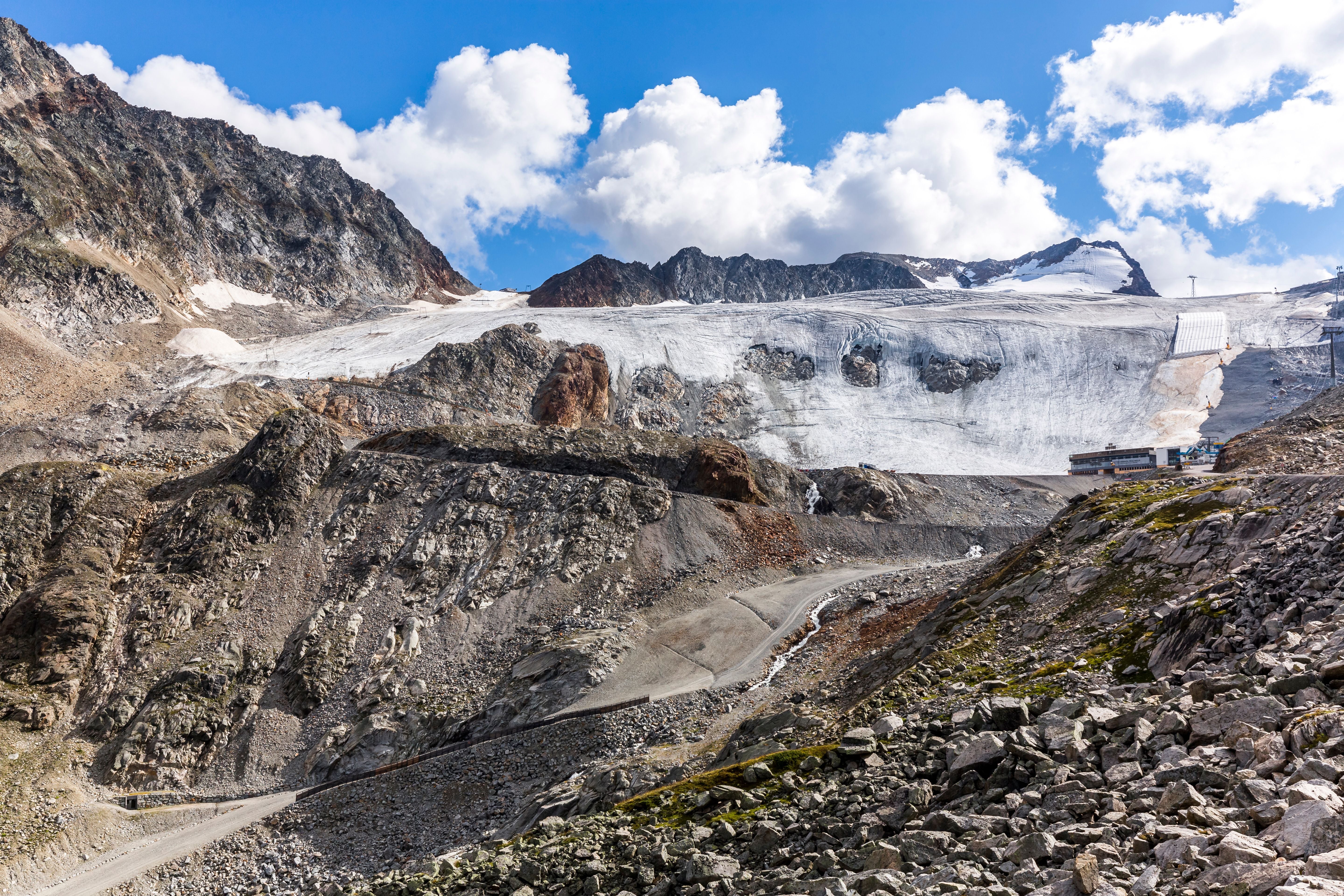 Gerölllandschaft vor einem schneebedeckten Gletscher mit blauem Himmel und Wolken.
