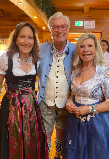 Drei Personen in traditioneller bayerischer Tracht lächeln in einem Holzraum in die Kamera.