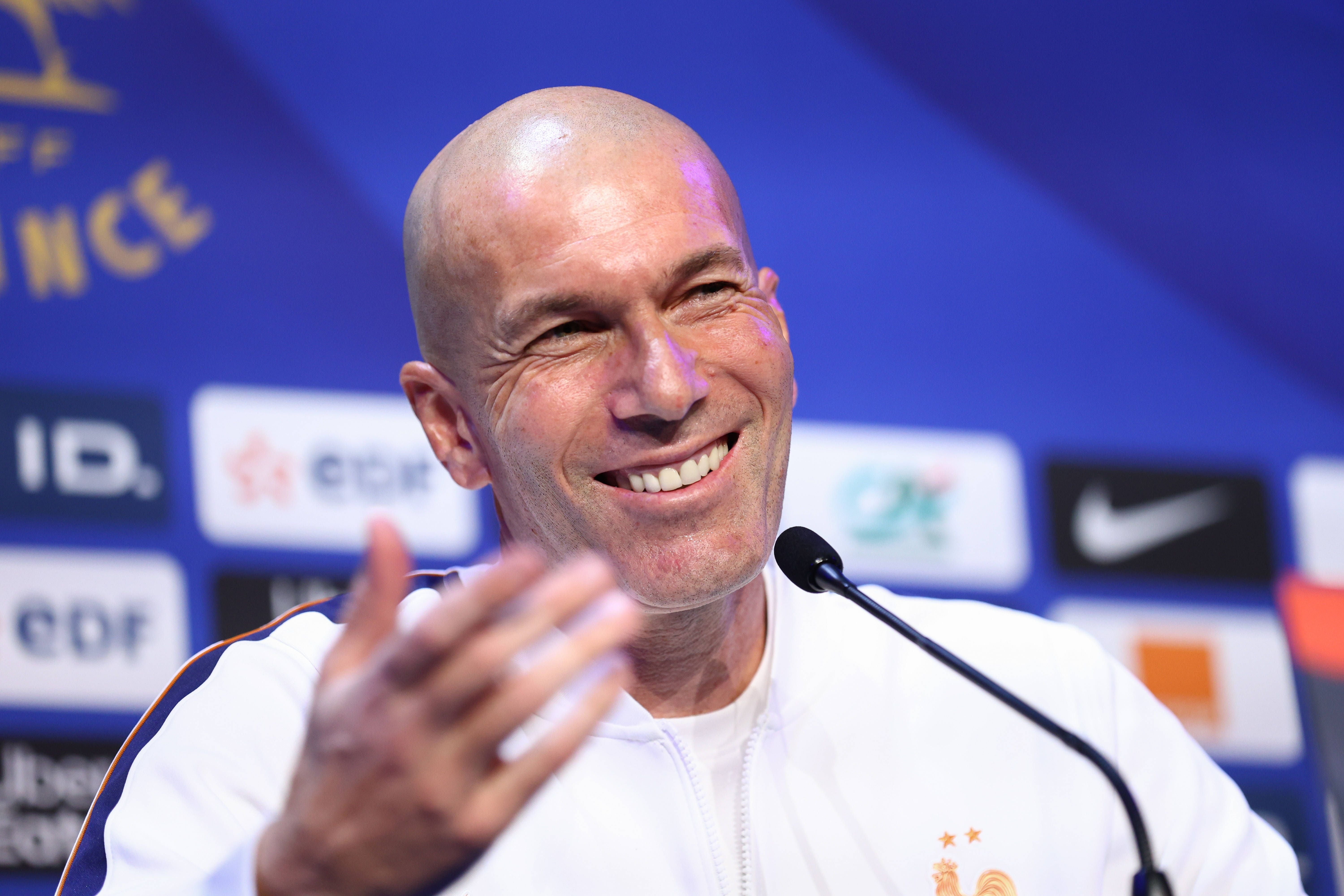 Zinedine Zidane lächelt auf einer Pressekonferenz vor einem Mikrofonsystem.