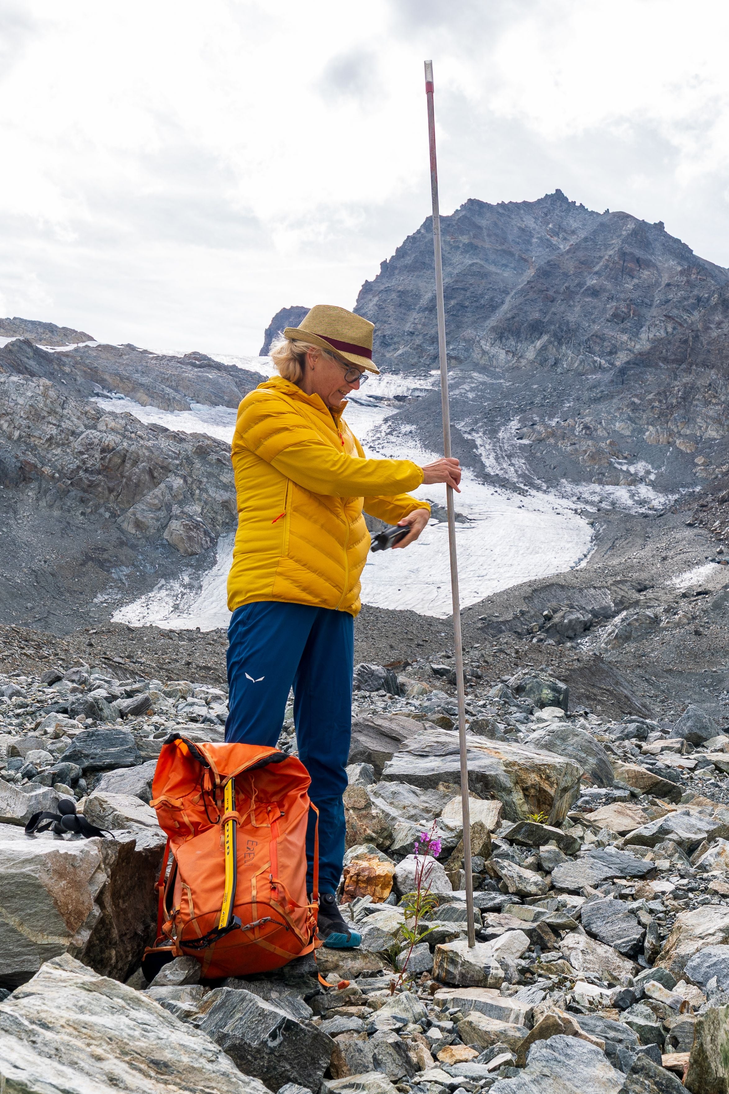 Eine Frau misst mit einem Stab im Geröll vor einem Gletscher in den Alpen.