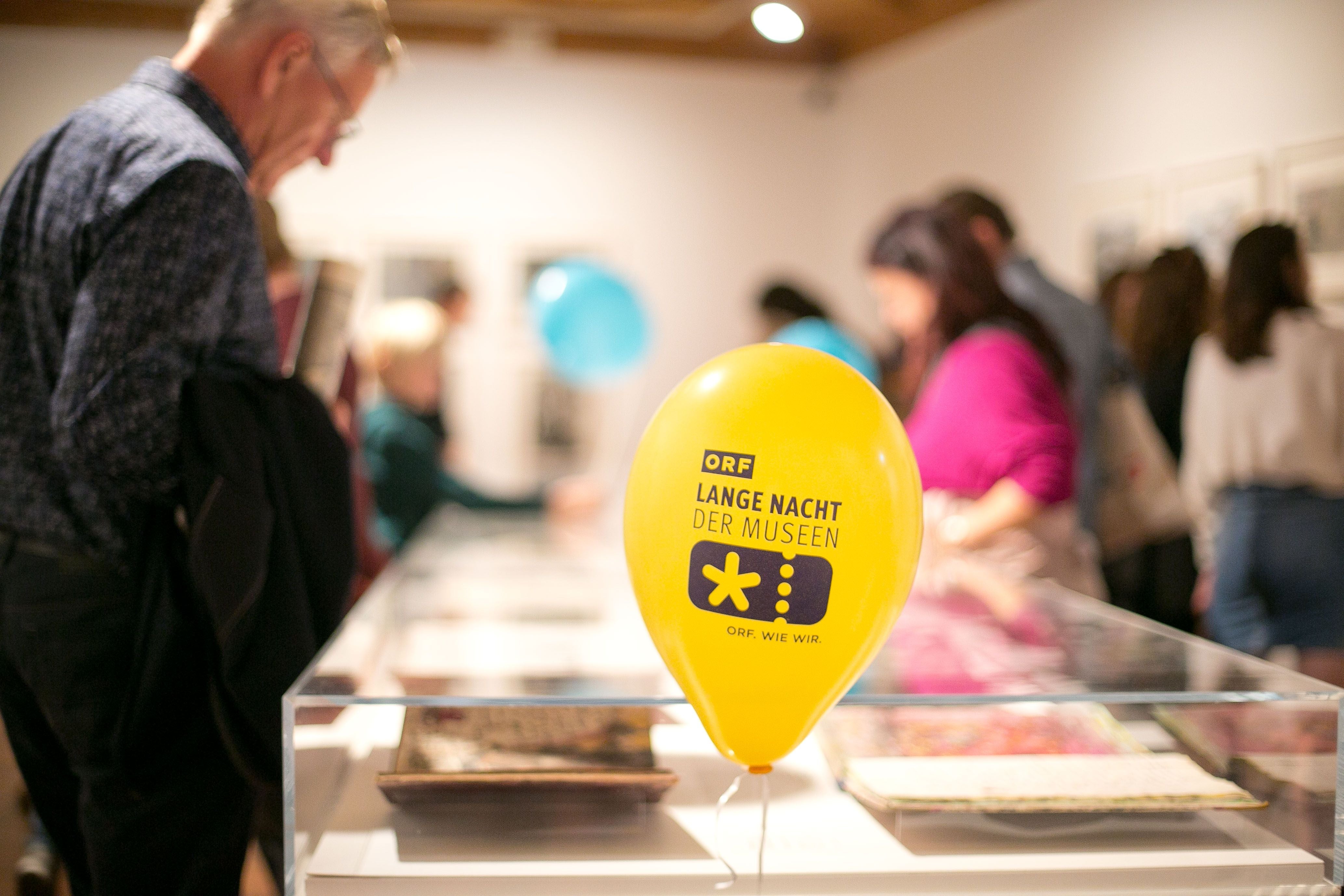 Gelber Luftballon mit "Lange Nacht der Museen" in einem Museum, Menschen im Hintergrund.