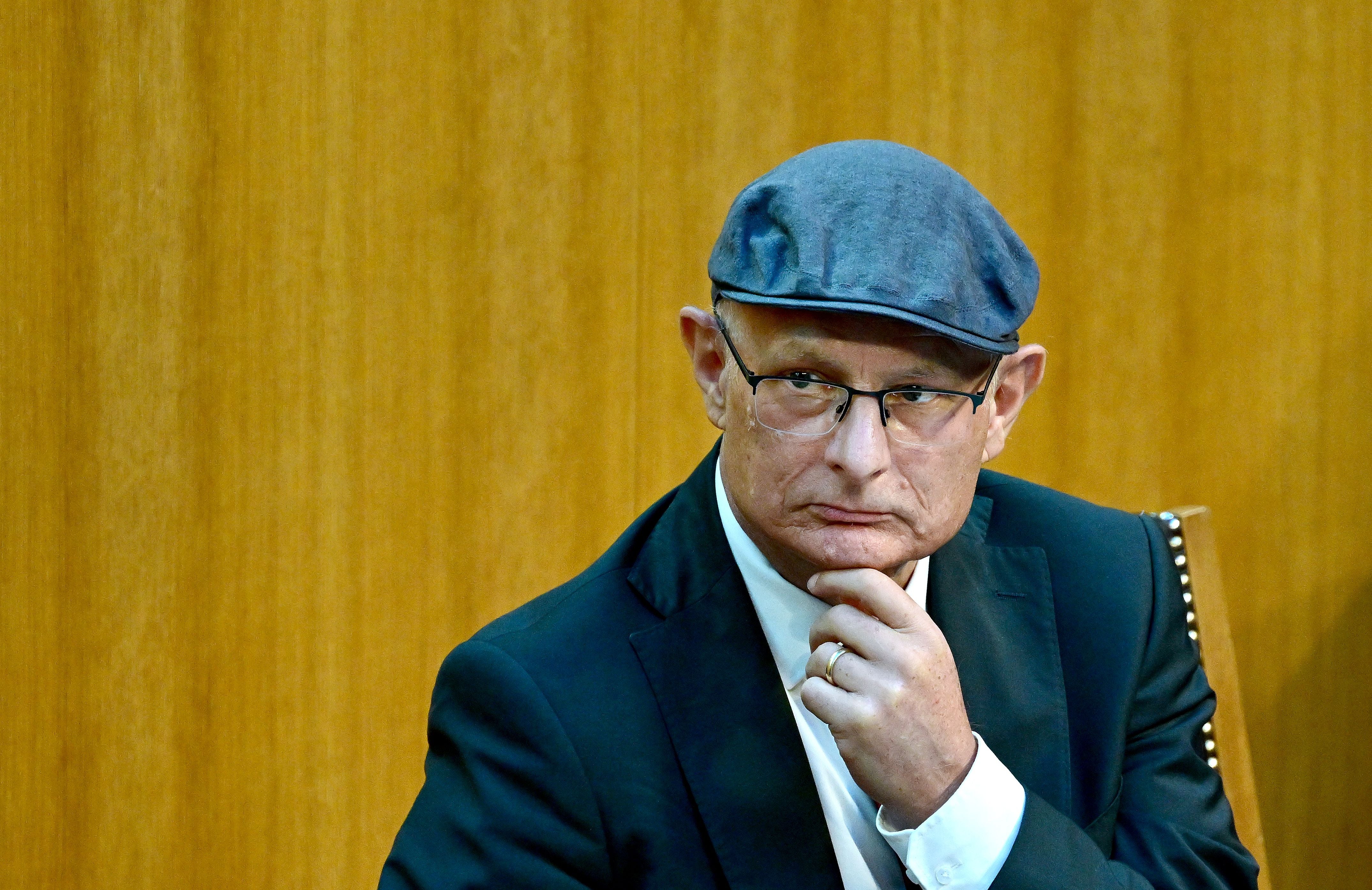 Ein Mann mit Brille und Mütze sitzt nachdenklich vor einer Holzwand.