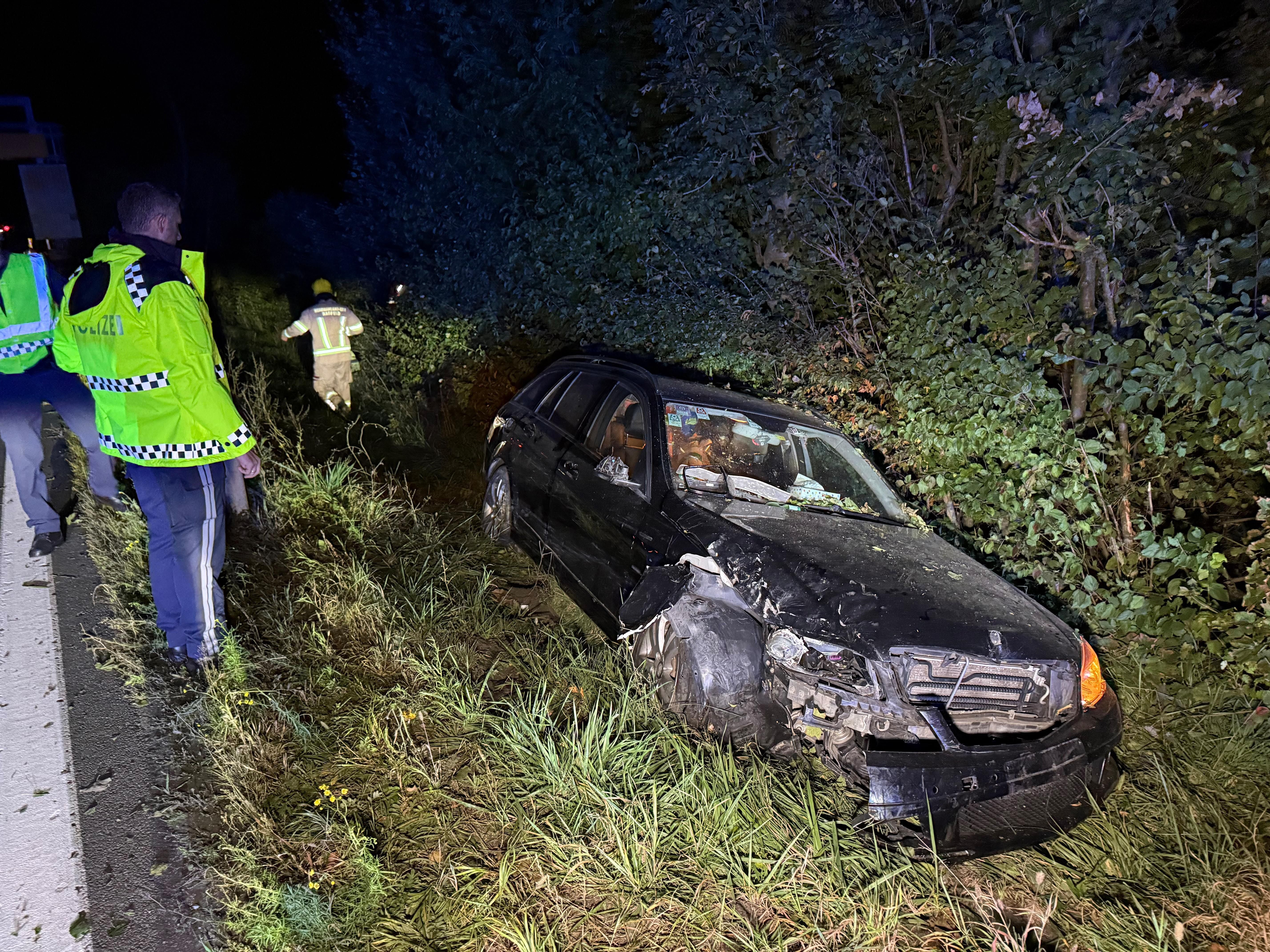 Polizisten und Feuerwehr inspizieren ein beschädigtes Auto im Straßengraben bei Nacht.