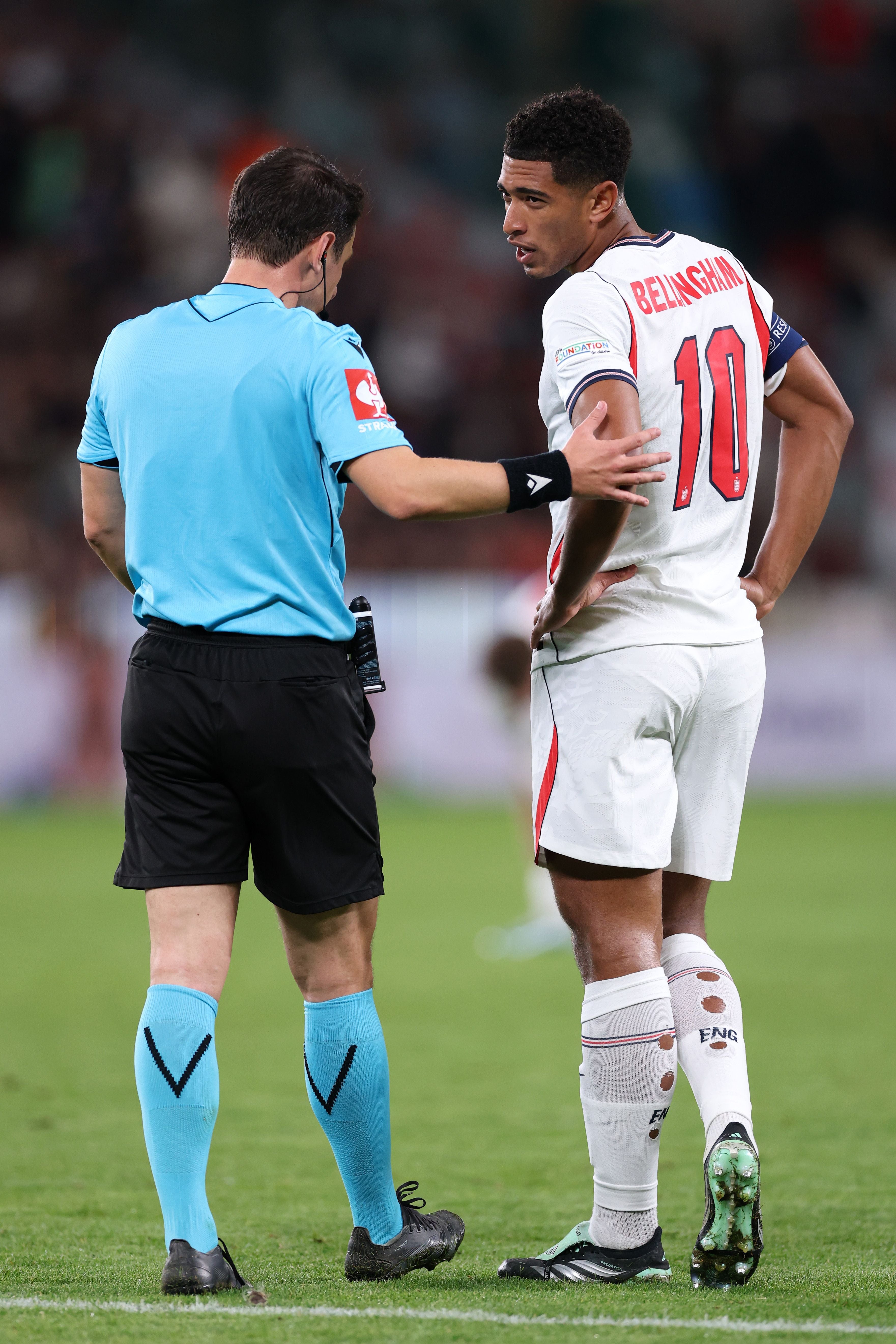 Jude Bellingham spricht im weißen England-Trikot mit Rückennummer 10 auf dem Spielfeld mit dem Schiedsrichter.