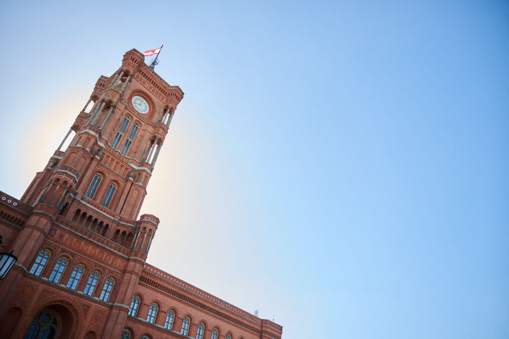 Das Rote Rathaus in Berlin mit Uhrturm und wehender Flagge vor blauem Himmel.