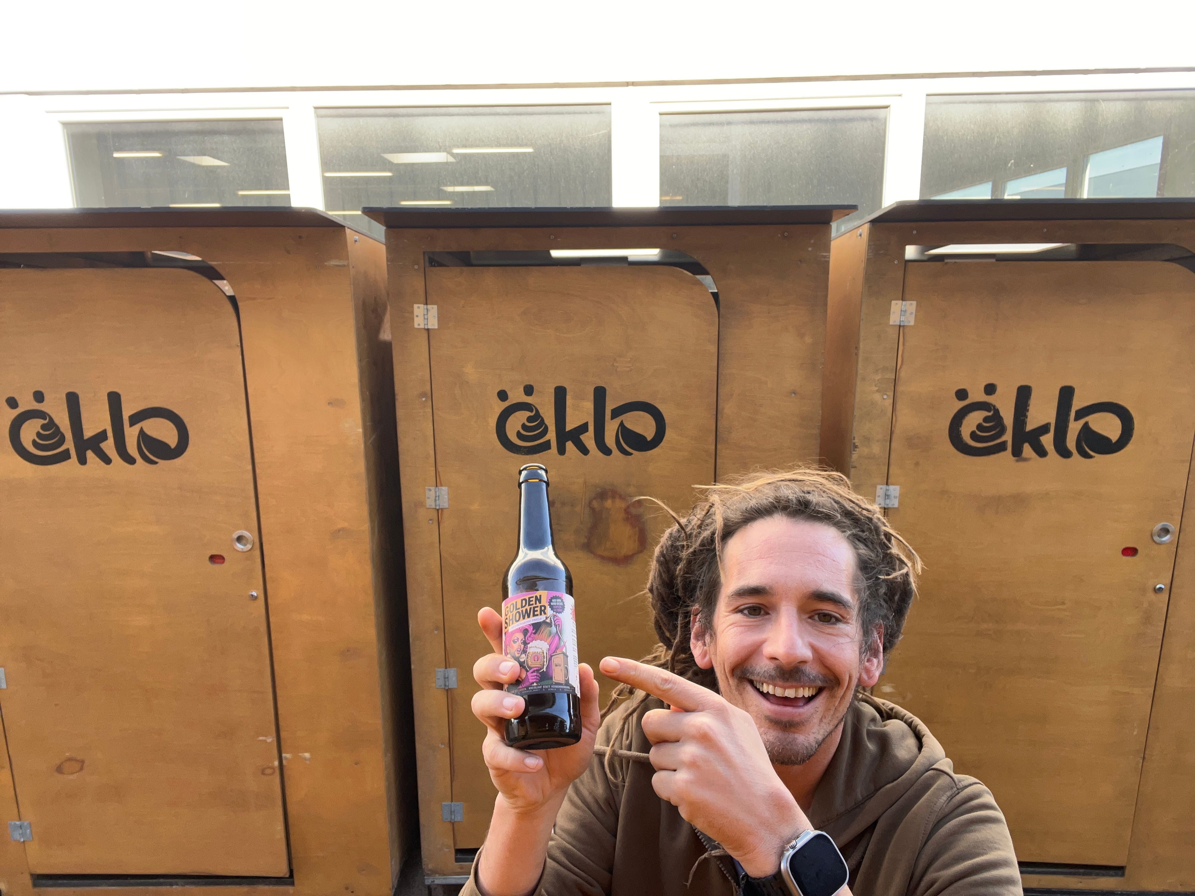 Lachender Mann hält eine Flasche Urin-Bier vor Komposttoiletten mit „öklo“-Logo.