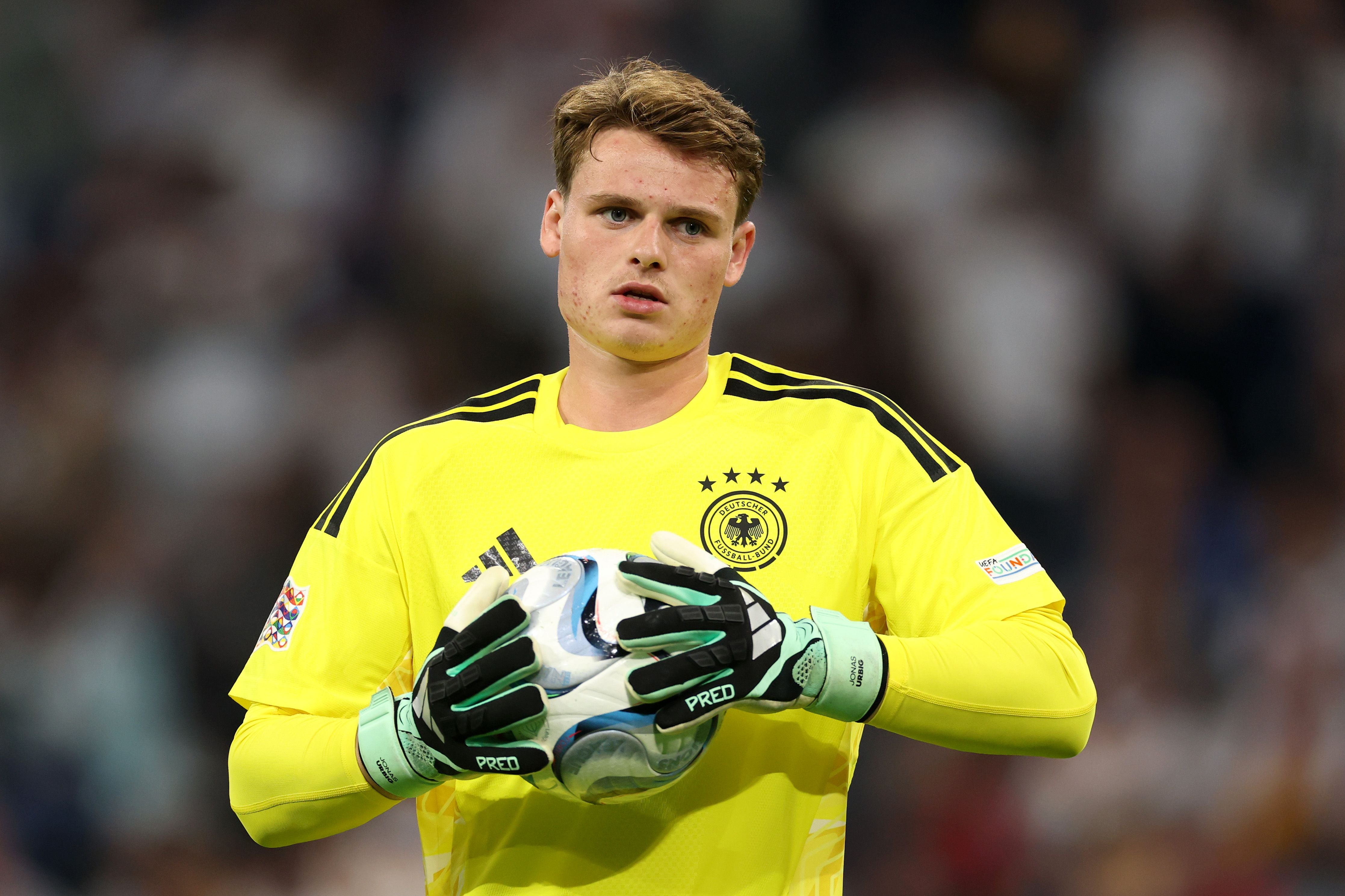 Ein Torwart im gelben Trikot hält einen Fußball bei einem Spiel in einem Stadion.