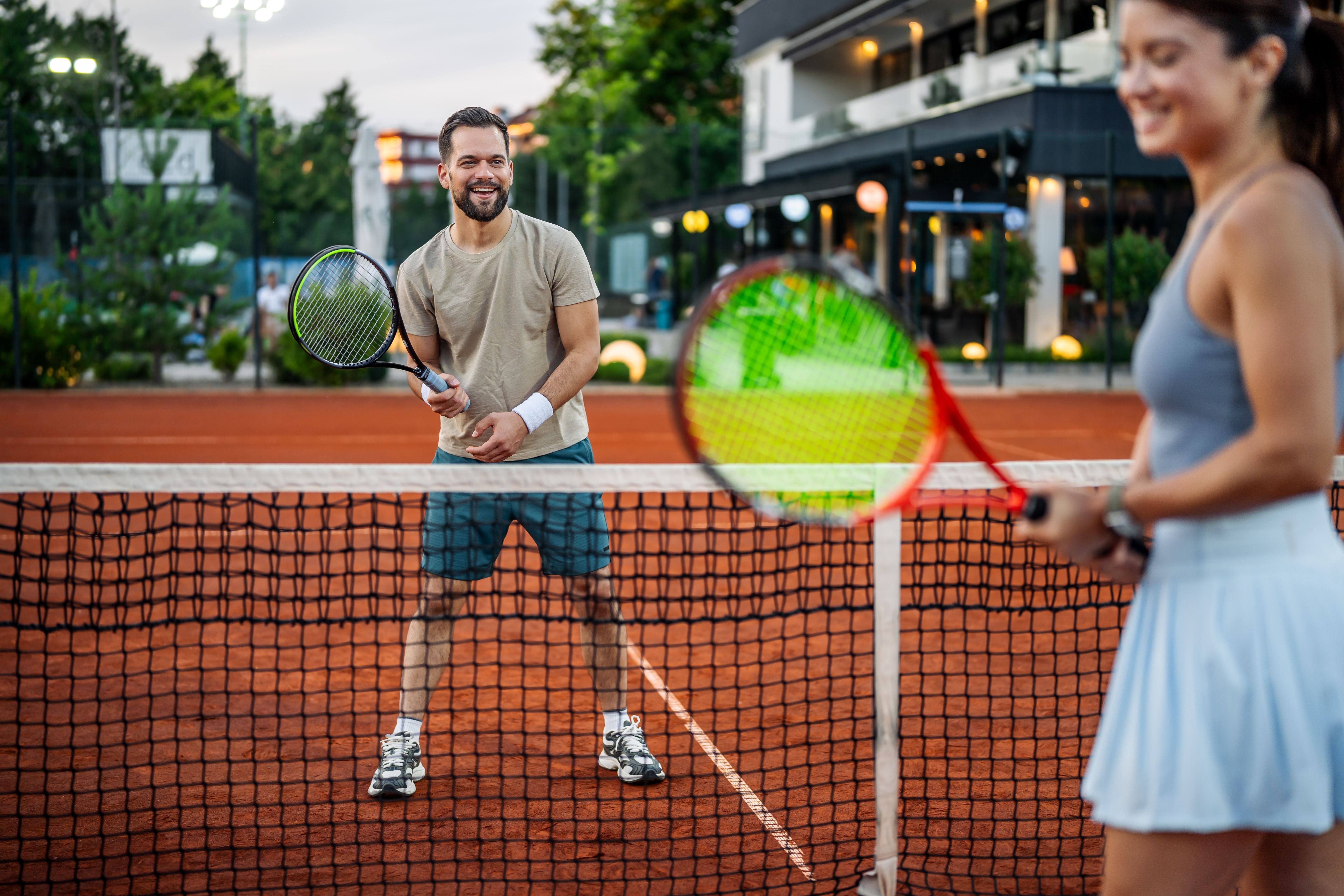 Junges Paar spielt lachend Tennis auf einem Sandplatz, beide halten Tennisschläger am Netz.