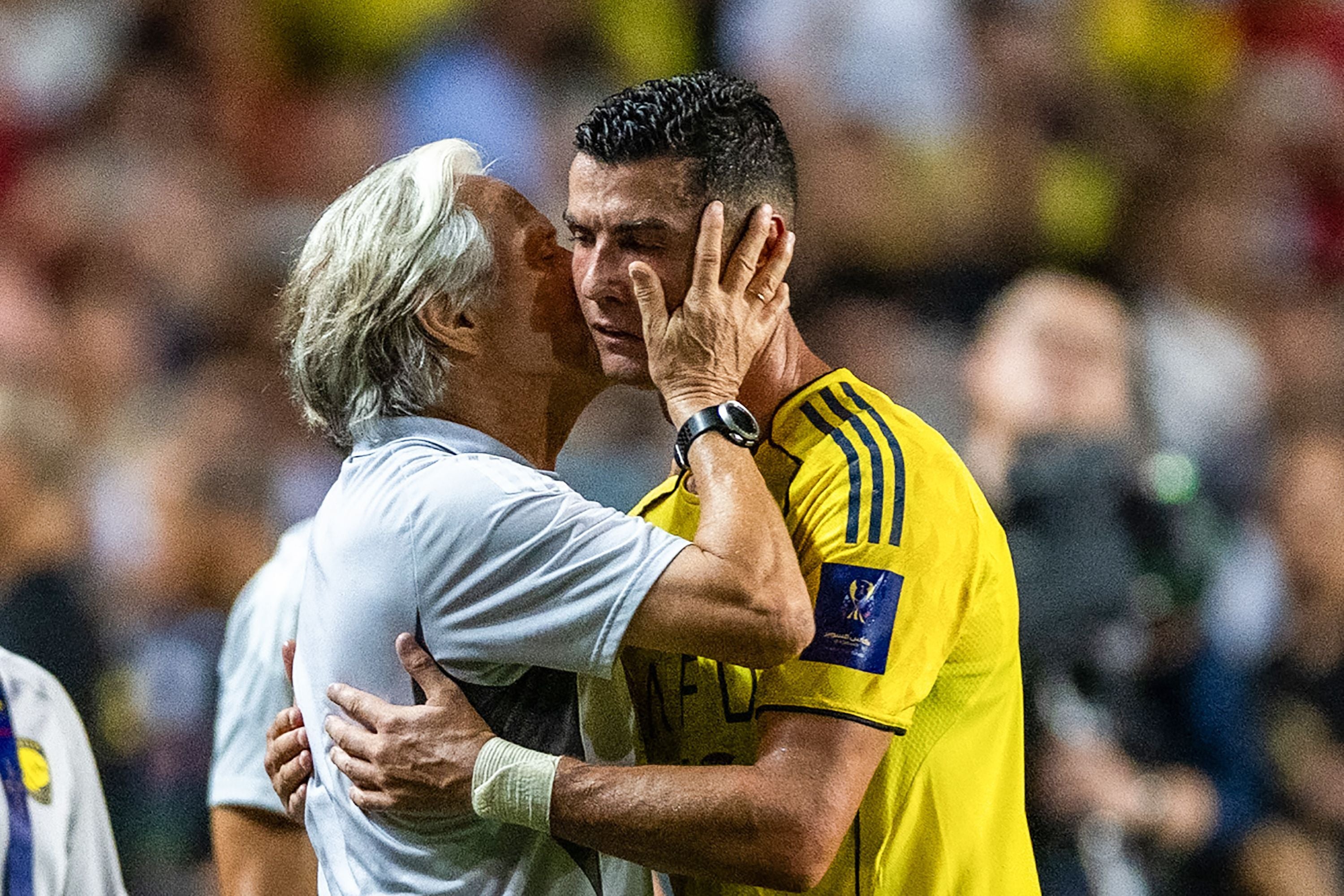 Trainer umarmt emotional einen Fußballspieler im gelben Trikot auf dem Spielfeld.