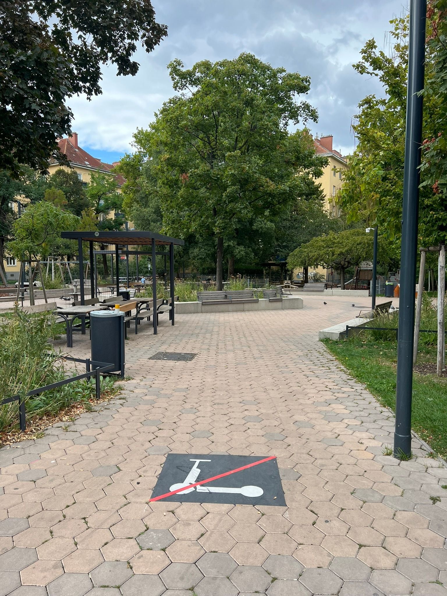 Weg im Park mit E-Scooter-Verbotsschild auf dem Boden, Bänke und Bäume im Hintergrund.