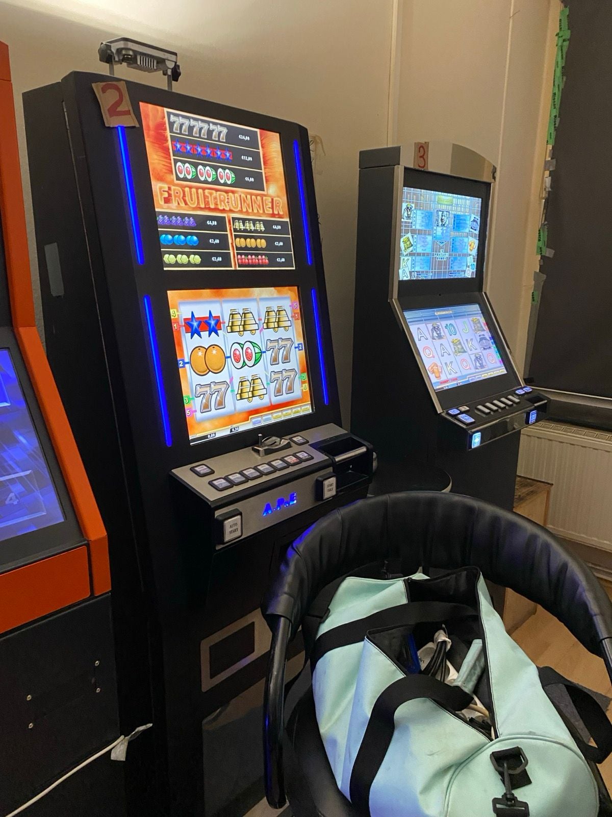 Zwei Spielautomaten in einem Raum, davor ein schwarzer Stuhl mit einer Sporttasche.