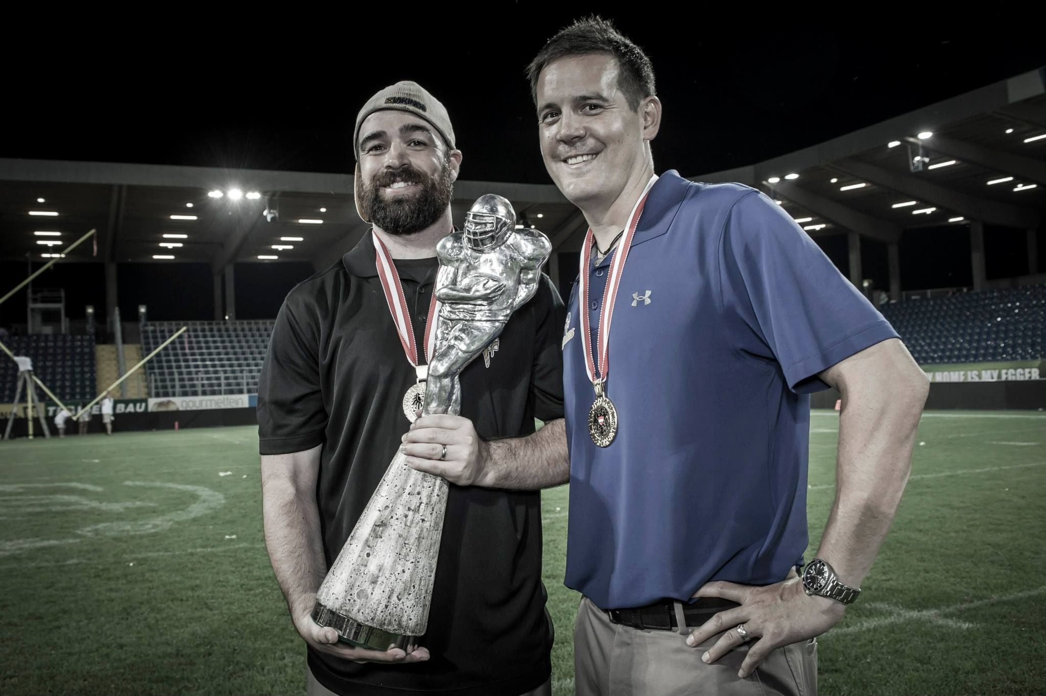 Zwei Männer mit Medaillen halten auf einem Footballfeld einen großen Pokal in Form eines Footballspielers.