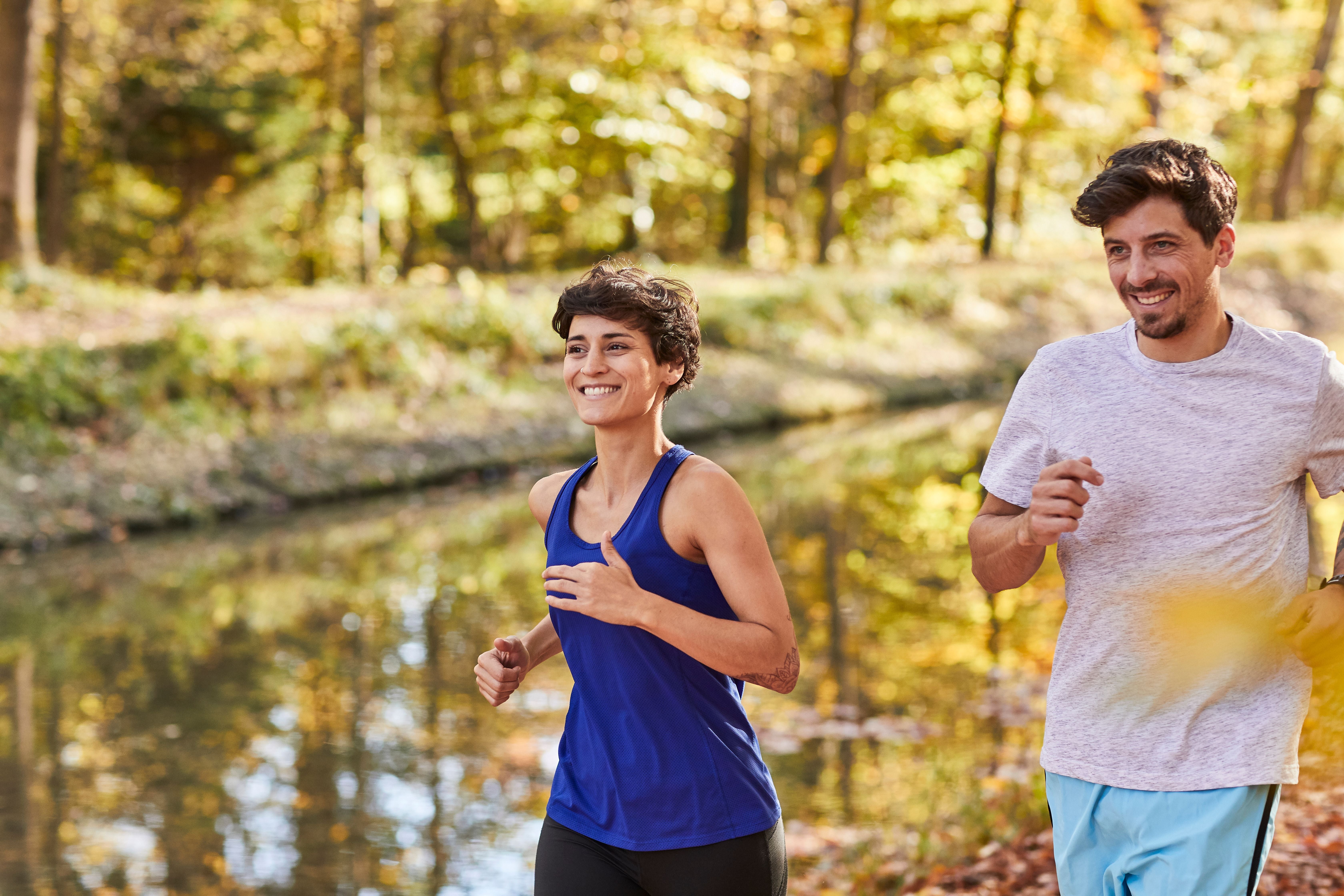 Zwei Menschen joggen lächelnd im Herbst an einem Flussufer entlang.