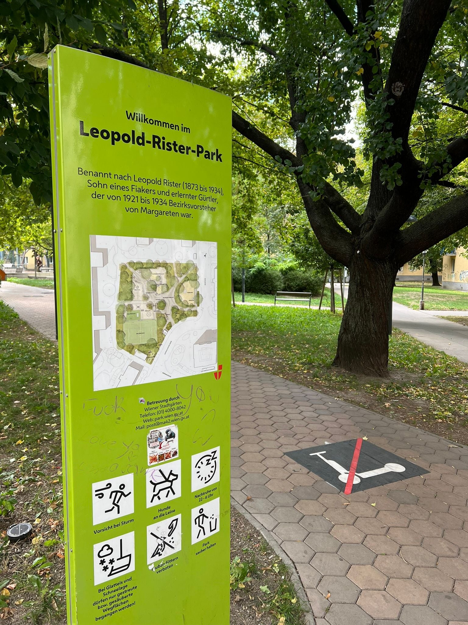 Grünes Infoschild zum Leopold-Rister-Park neben einem Baum und einem Parkweg mit Verbotsschild für E-Scooter.