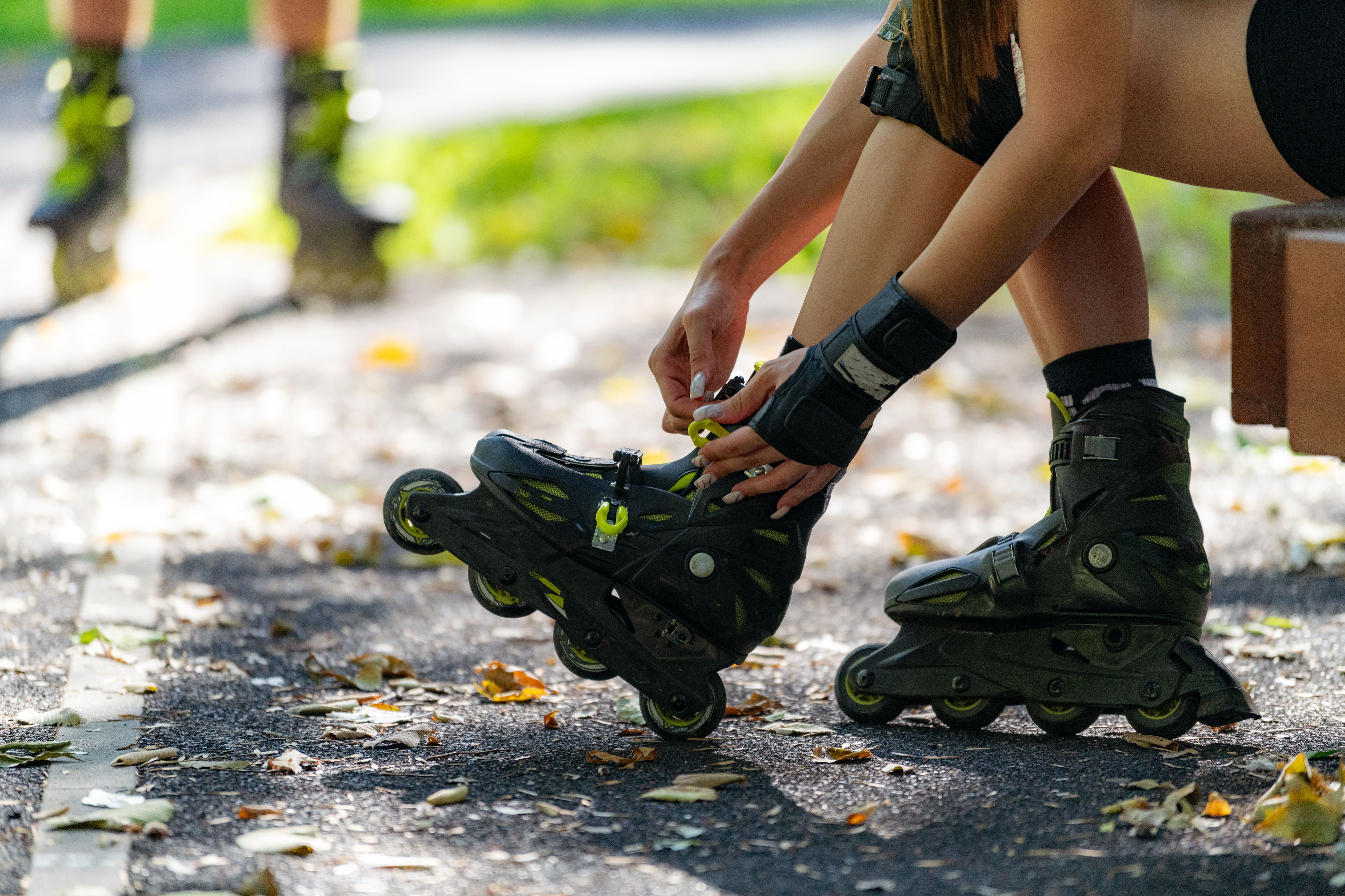 Eine Person zieht draußen im Park Inlineskates an, im Hintergrund steht eine weitere Person.