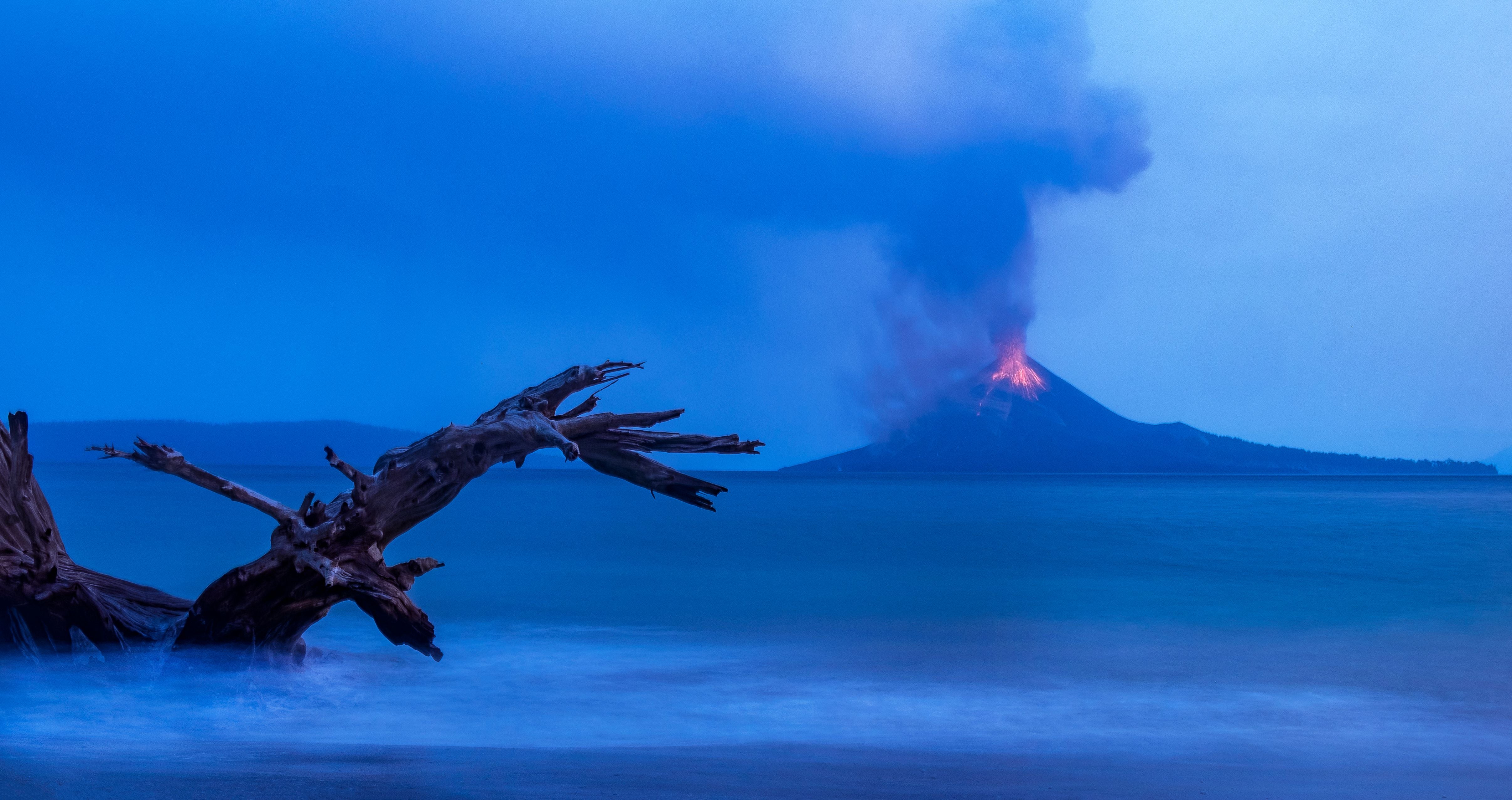 Vulkan Anak Krakatau stößt Rauch und Lava aus, im Vordergrund ein Treibholz am Strand.