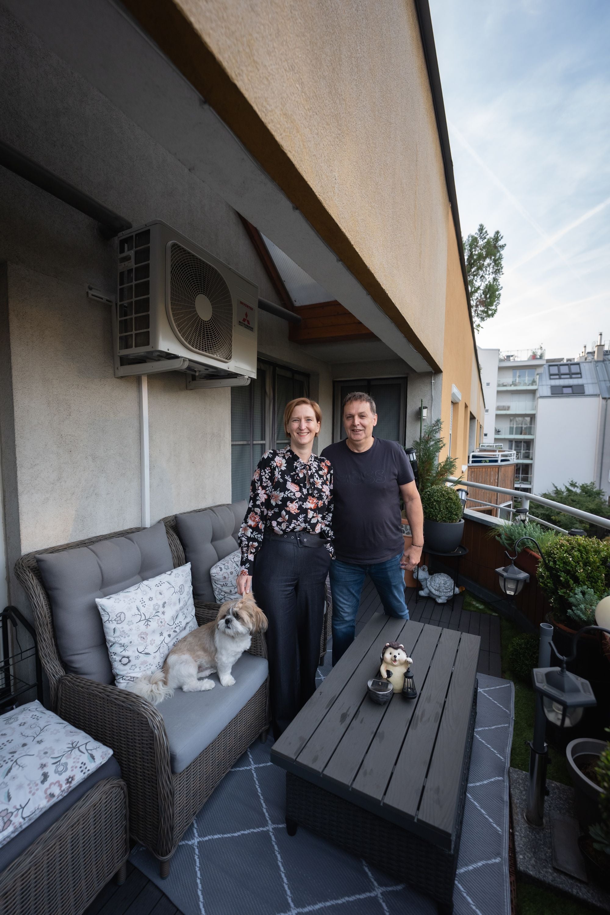 Eine Frau und ein Mann stehen auf einem Balkon mit Sitzbank, Hund und Klimaanlage an der Wand.