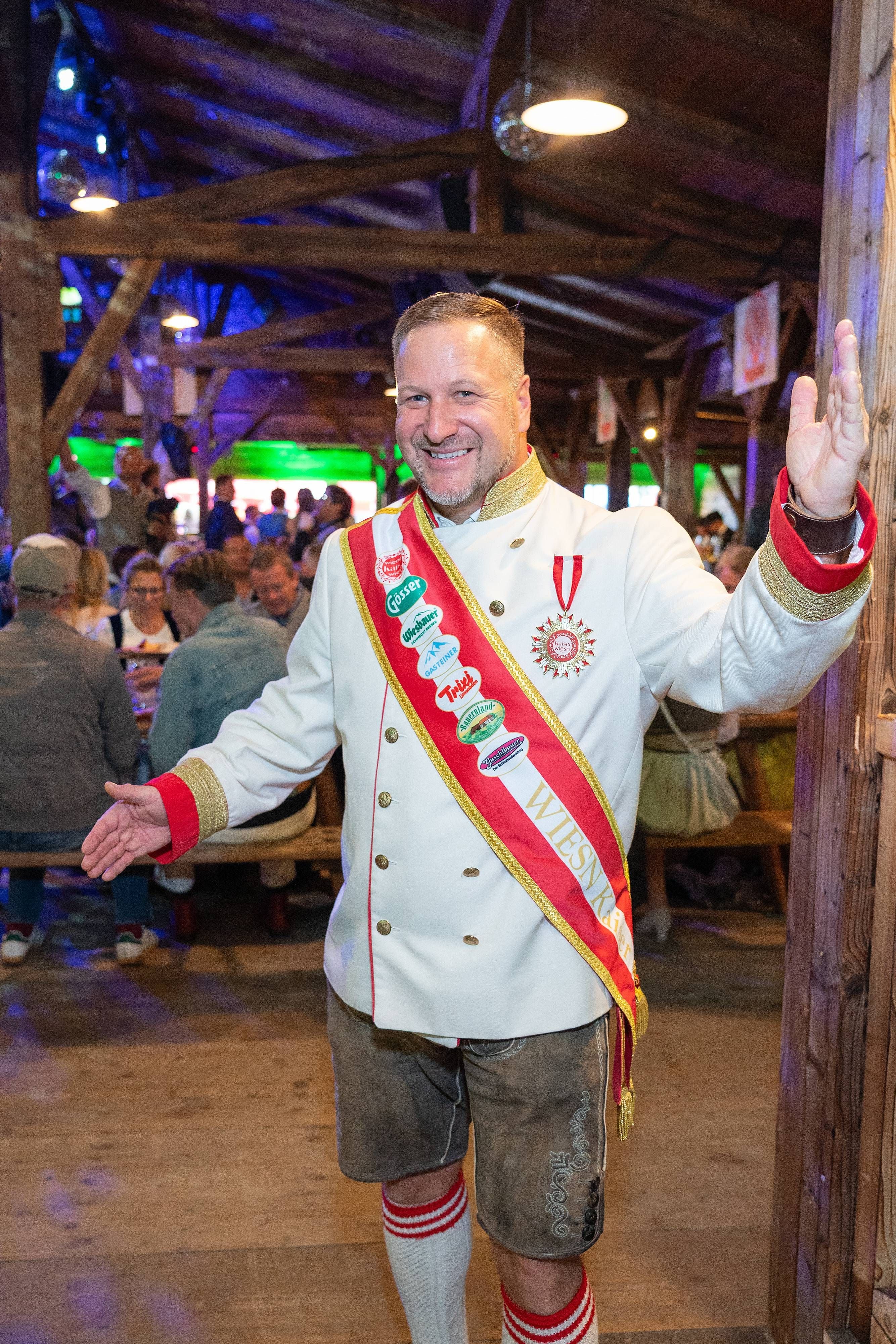 Ein Mann in Tracht mit Schärpe und Orden begrüßt Gäste auf einem Fest in einer Holzhütte.