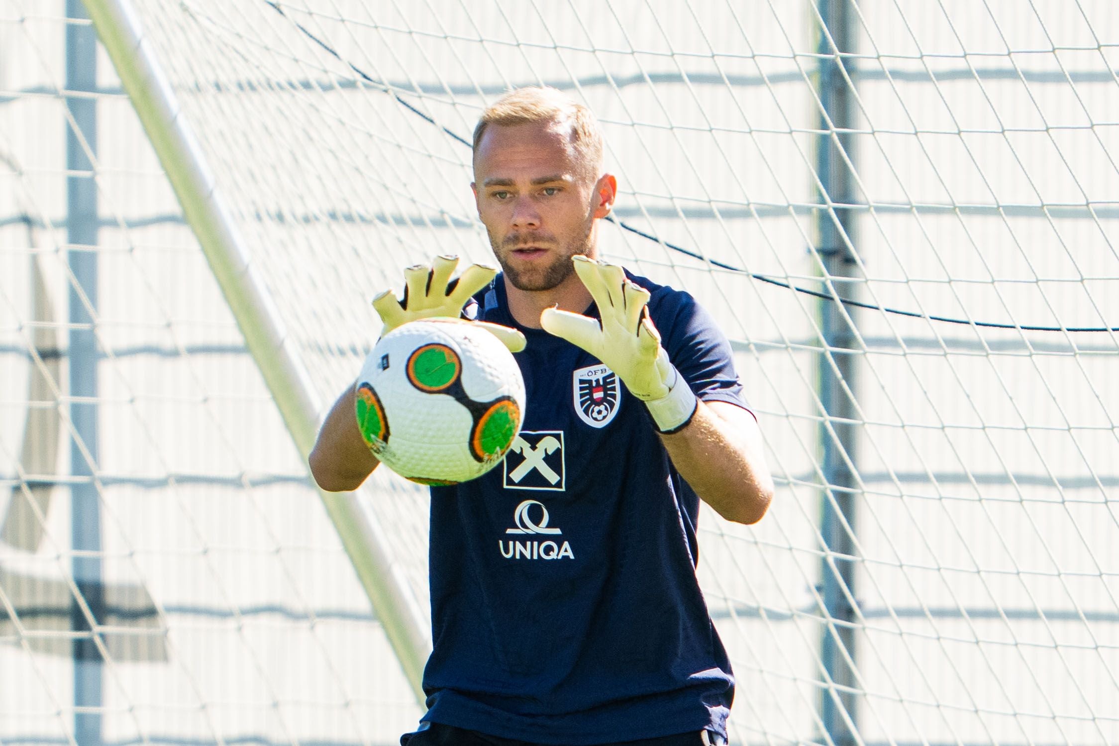 Ein Torwart im blauen Trikot fängt beim Training einen Fußball vor dem Tor ab.