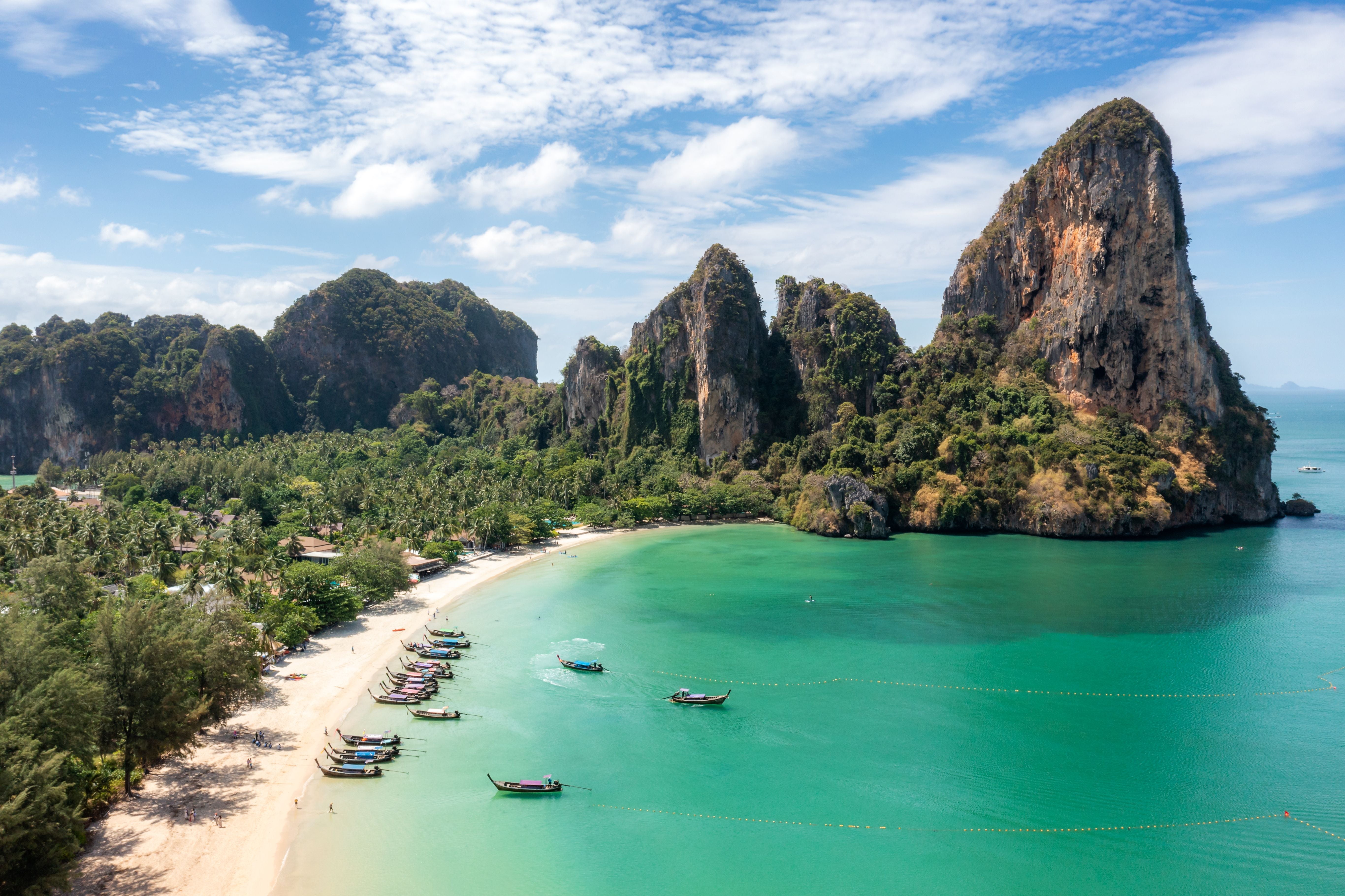 Aerialaufnahme von Railay Beach mit türkisfarbenem Meer, langen Booten und grünen Kalksteinfelsen.