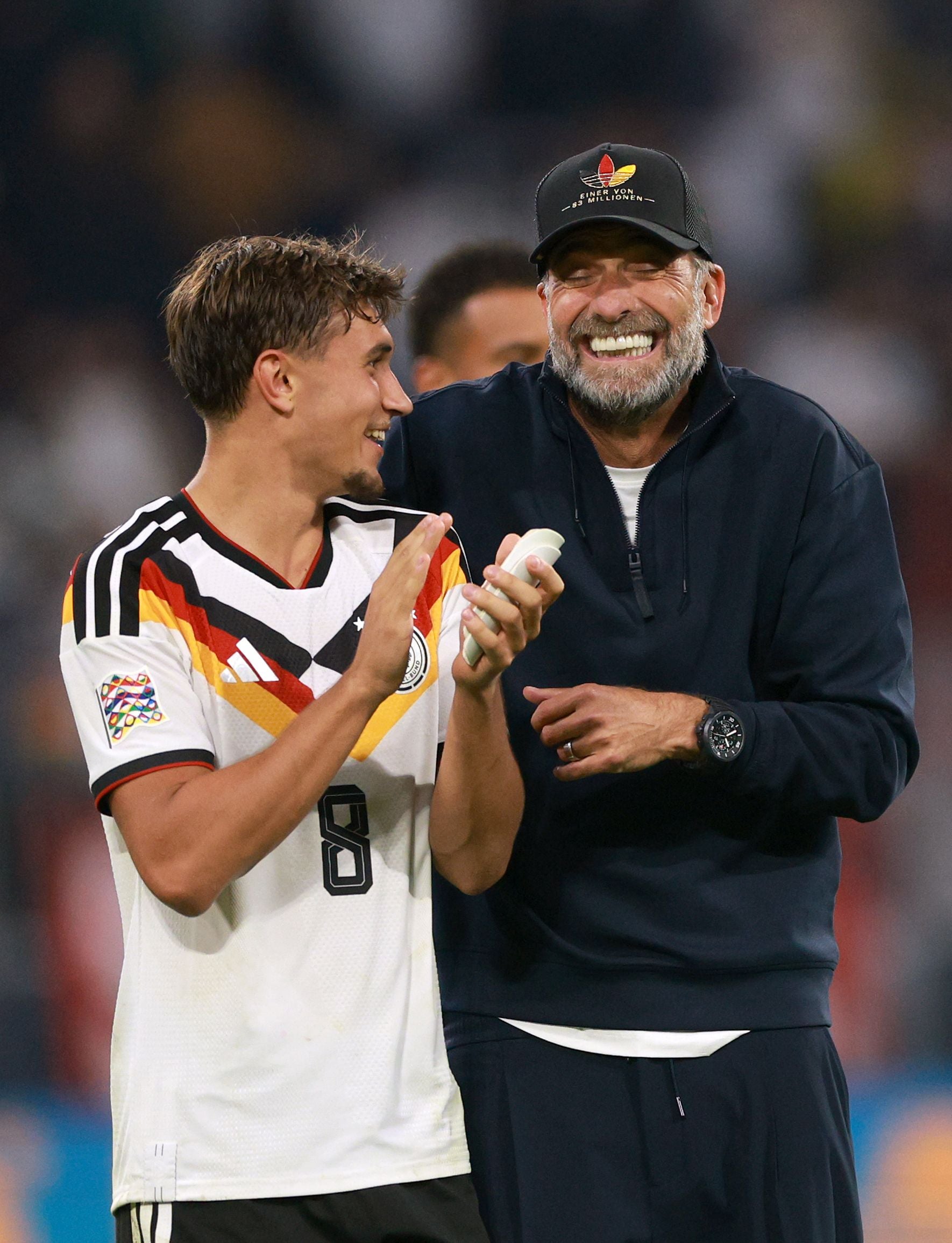 Zwei Männer lachen und unterhalten sich auf einem Fußballfeld, einer trägt ein Deutschland-Trikot.