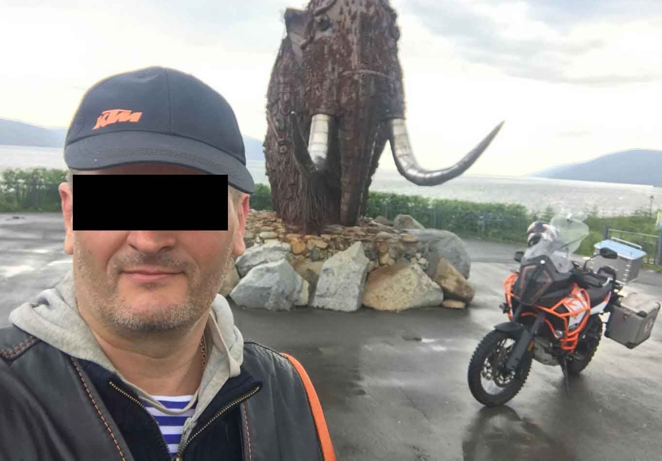 Mann mit schwarzer Kappe vor Mammutstatue und Motorrad auf nasser Straße.