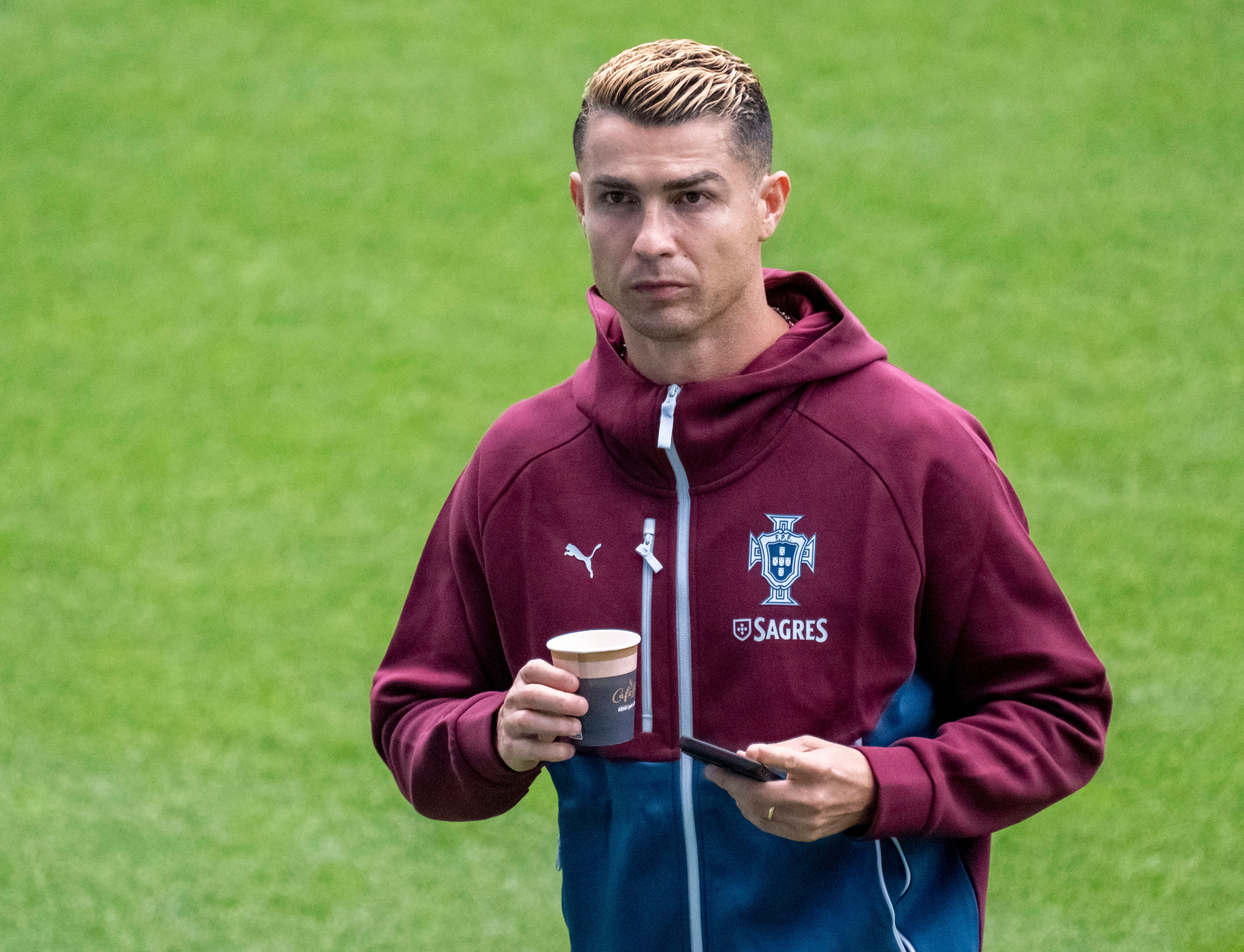 Ein Fußballspieler im Trainingsanzug hält einen Kaffeebecher und ein Handy auf einem Sportfeld.