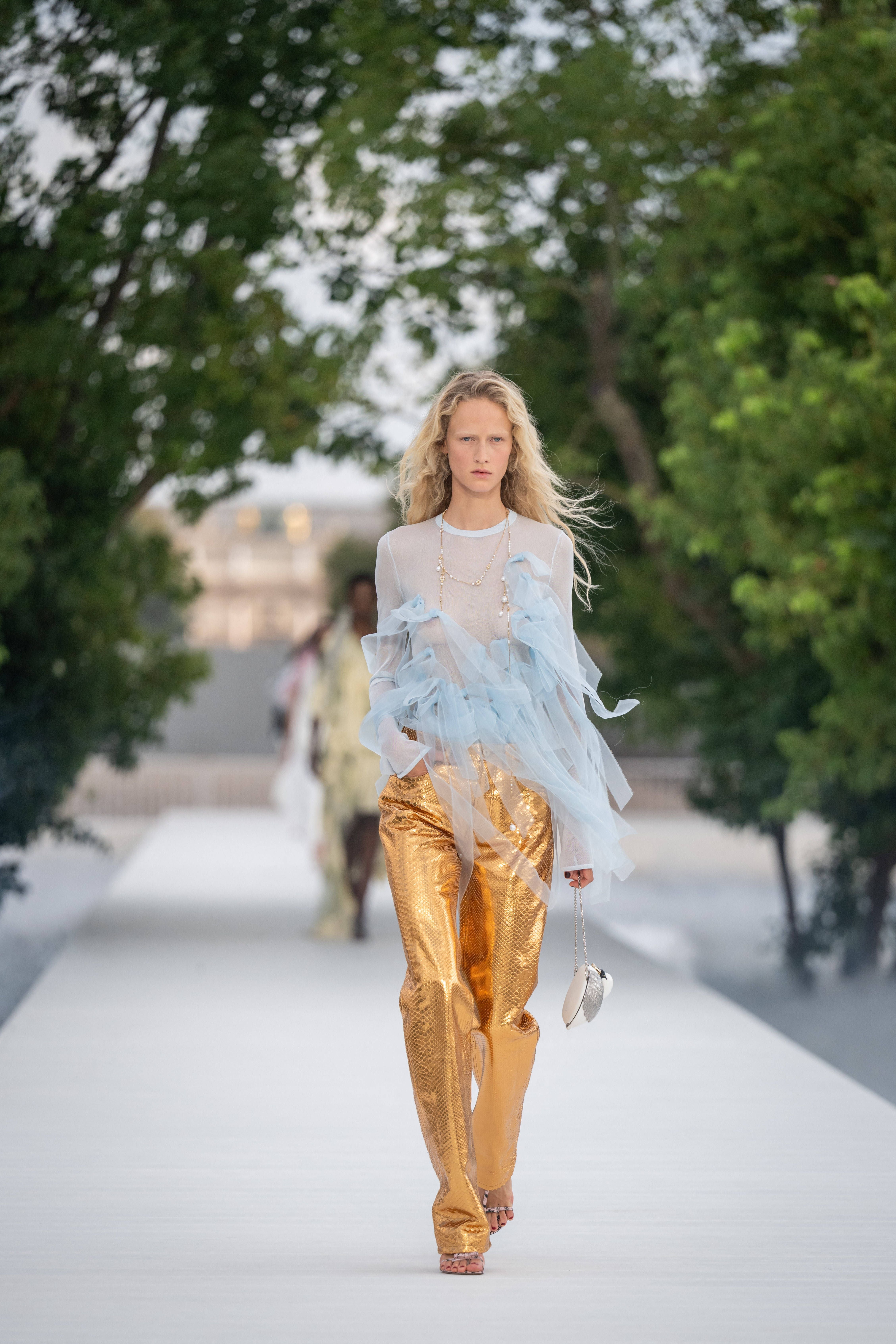 Model in goldener Hose und transparentem Oberteil mit blauem Tüll auf dem Dior-Laufsteg im Freien.