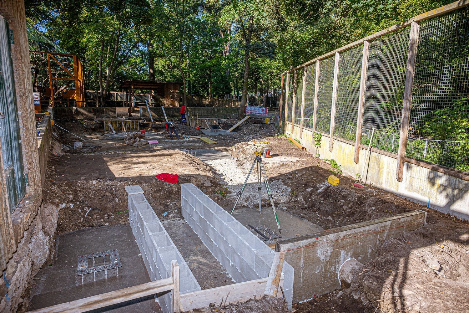Baustelle im Tiergarten mit Mauerbau und Baugeräten, umgeben von Bäumen und Zäunen.